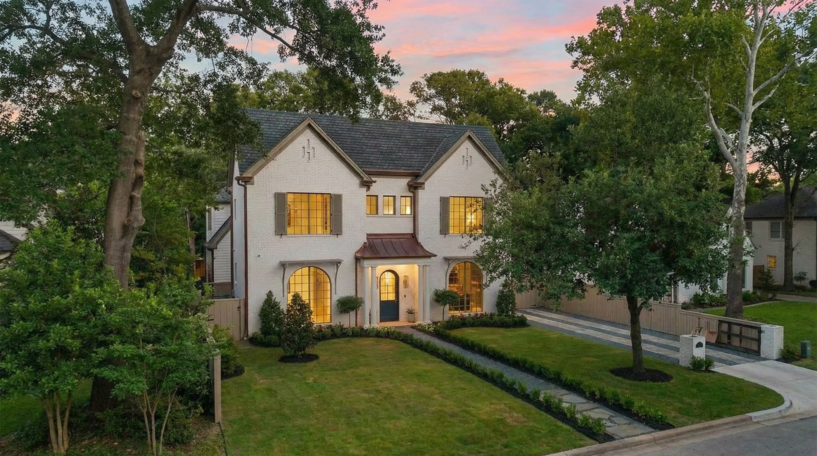 This image showcases the front exterior of a beautiful two-story home. The facade features a white brick exterior, a gabled roof with dark shingles, and arched windows, giving it a charming and refined appearance. Mature trees surround the property, adding to its curb appeal and creating a sense of privacy.
