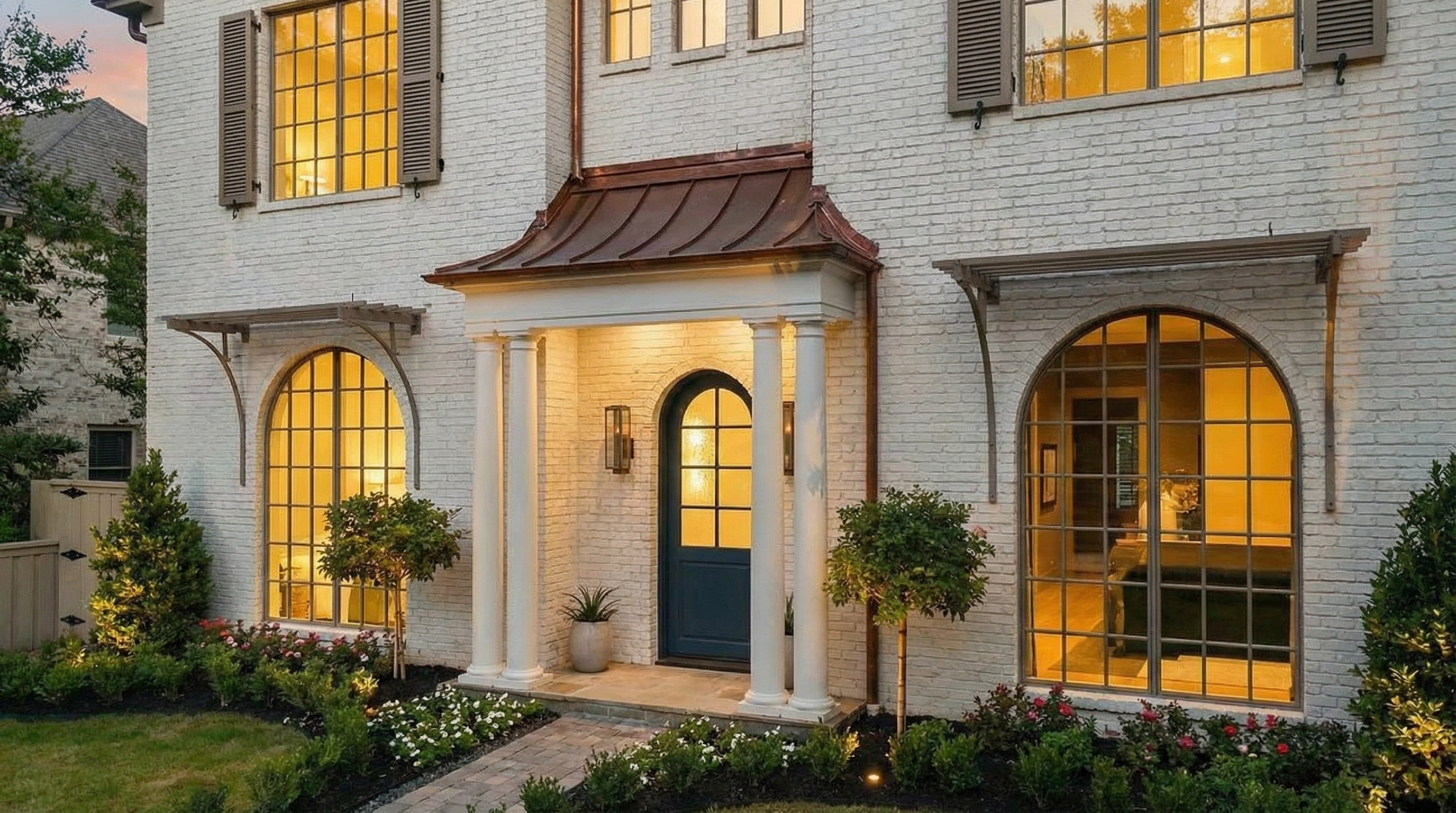 This is a lovely front view of a brick house. It shows well-maintained landscaping, a welcoming entry with columns and arched windows. The warm lighting emanating from the windows creates an inviting appearance.