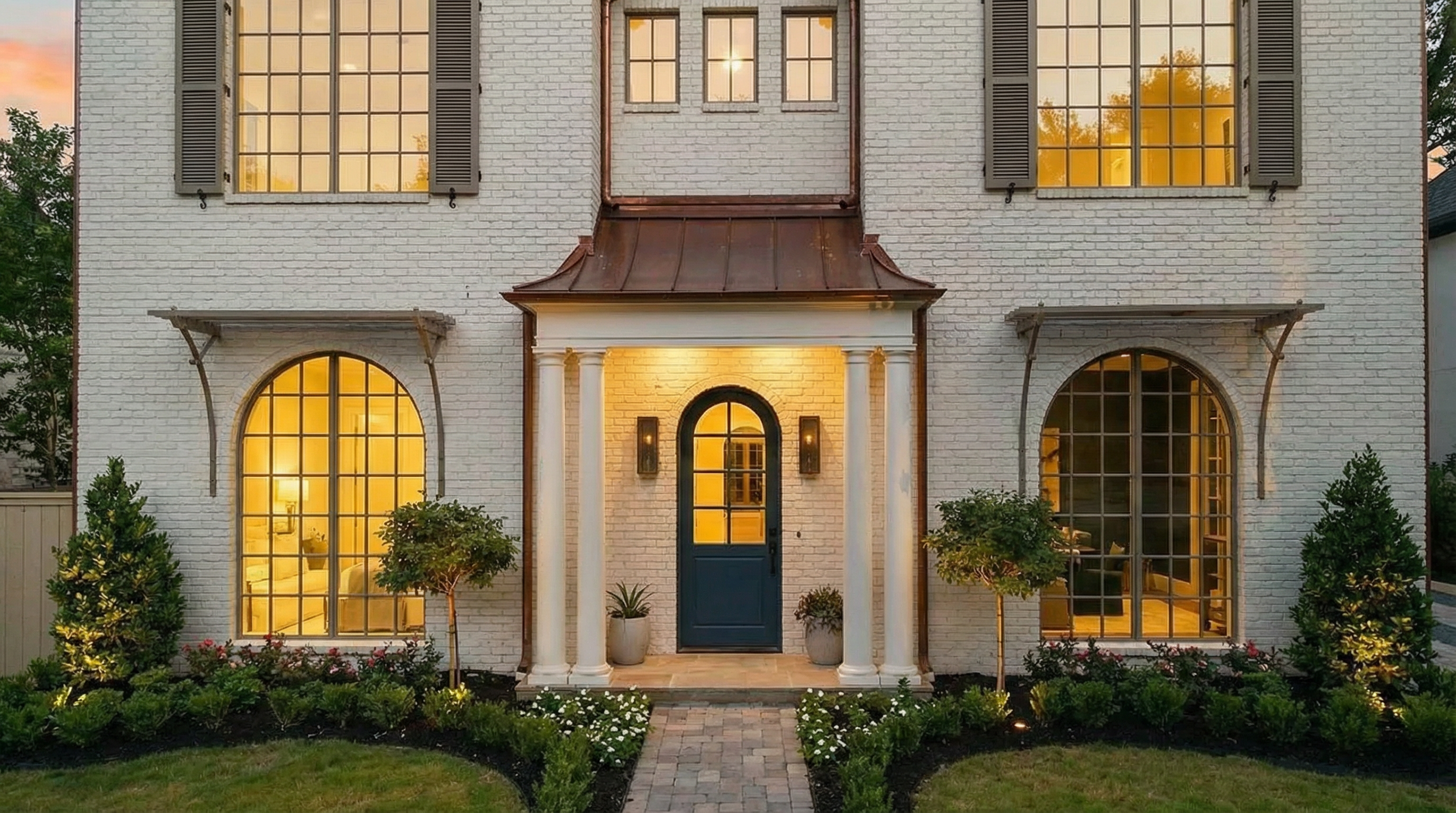 This is a front view of a beautifully designed home with a painted brick facade. The entryway features a stylish blue door framed by white columns and a copper-toned roof. Large windows with dark shutters add to the home's curb appeal, and the well-maintained landscaping enhances its inviting appearance.
