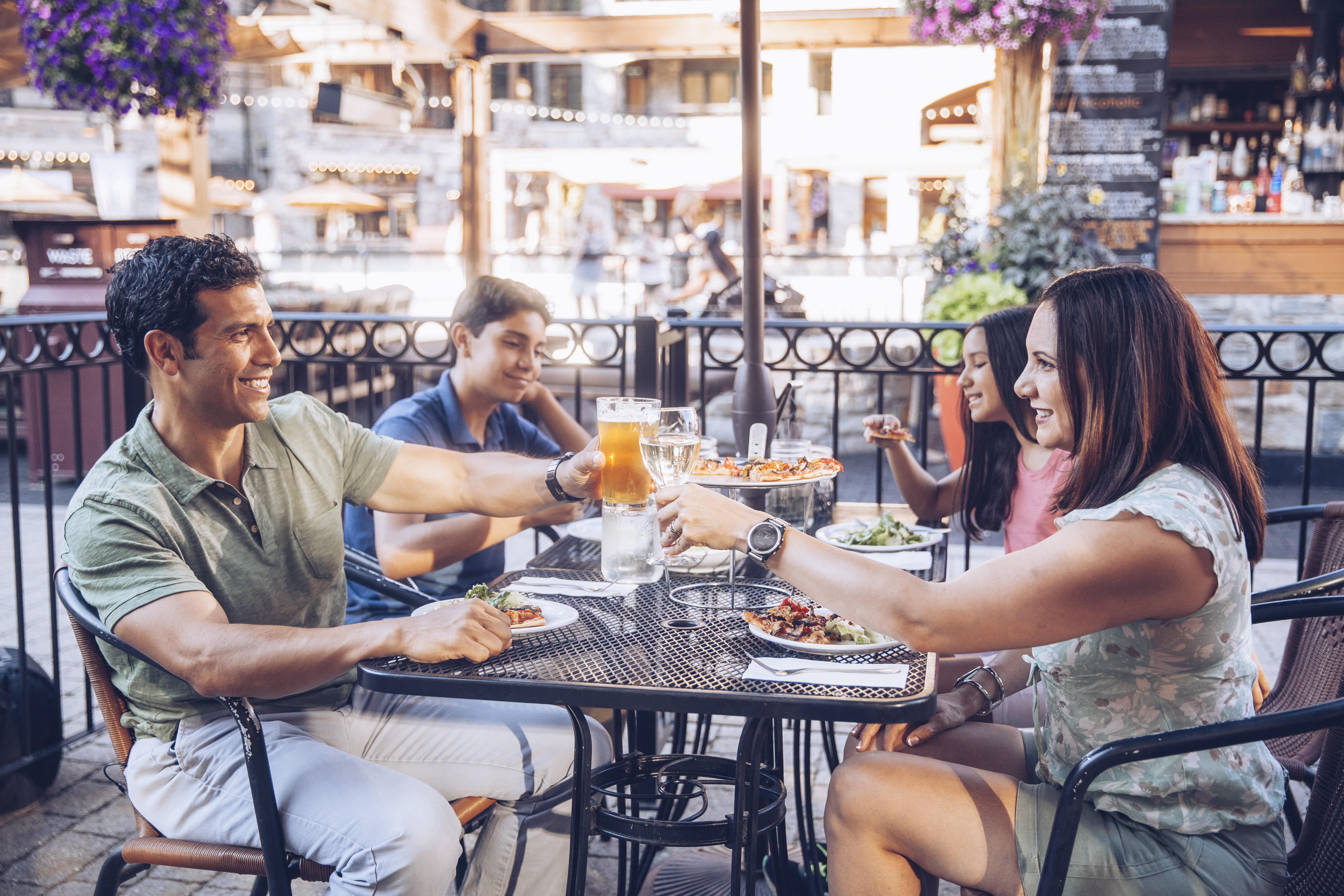 This image captures a family enjoying an outdoor dining experience on a patio, highlighting the property's inviting lifestyle amenities. The scene features a metal bistro table set with food and drinks, surrounded by a decorative metal railing and lush hanging floral baskets. The warm, golden-hour lighting creates a welcoming atmosphere, suggesting a vibrant community or restaurant setting perfect for social gatherings.