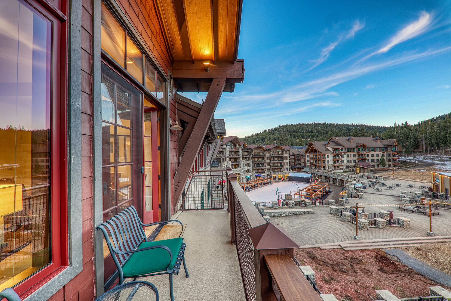 This elevated balcony offers a scenic vantage point overlooking a mountain resort village, complete with a central ice skating rink and surrounding lodge-style architecture. The balcony itself features a metal-framed bench with a green cushion, warm overhead lighting, and a sturdy railing, creating a cozy spot to enjoy the crisp mountain air. The perspective captures the transition from the private outdoor living space to the expansive, bustling community area below.