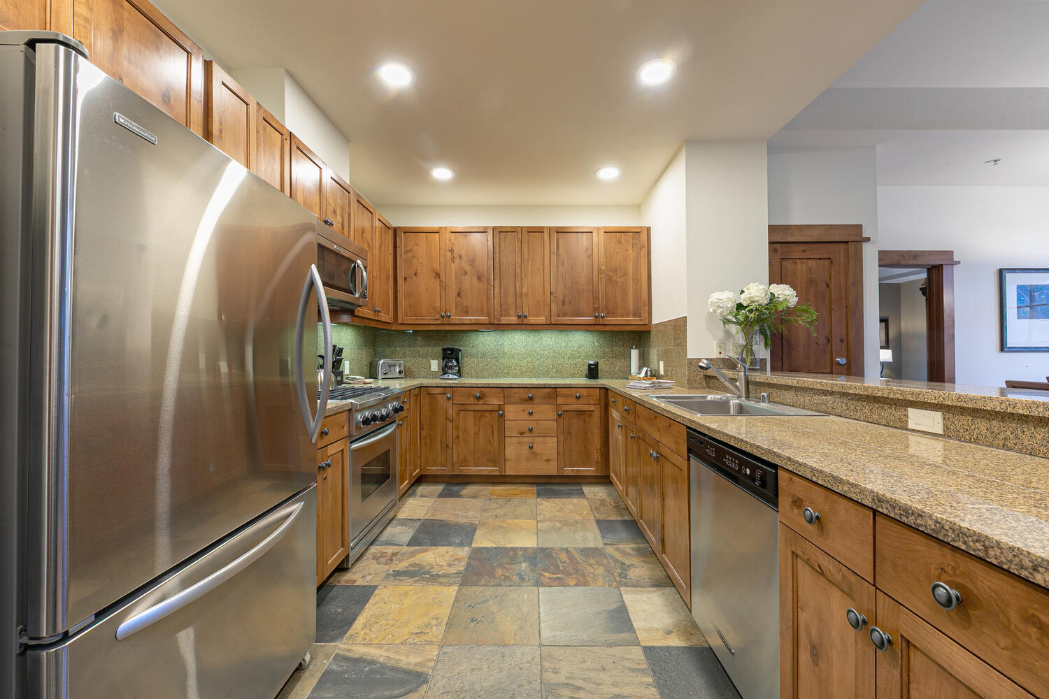 This U-shaped kitchen features warm-toned wood cabinetry paired with speckled stone countertops and a multi-colored slate tile floor. The space is equipped with stainless steel appliances, including a refrigerator, dishwasher, and range, and is illuminated by recessed ceiling lighting. A breakfast bar peninsula with a sink overlooks an adjacent living area, creating an open and functional layout.