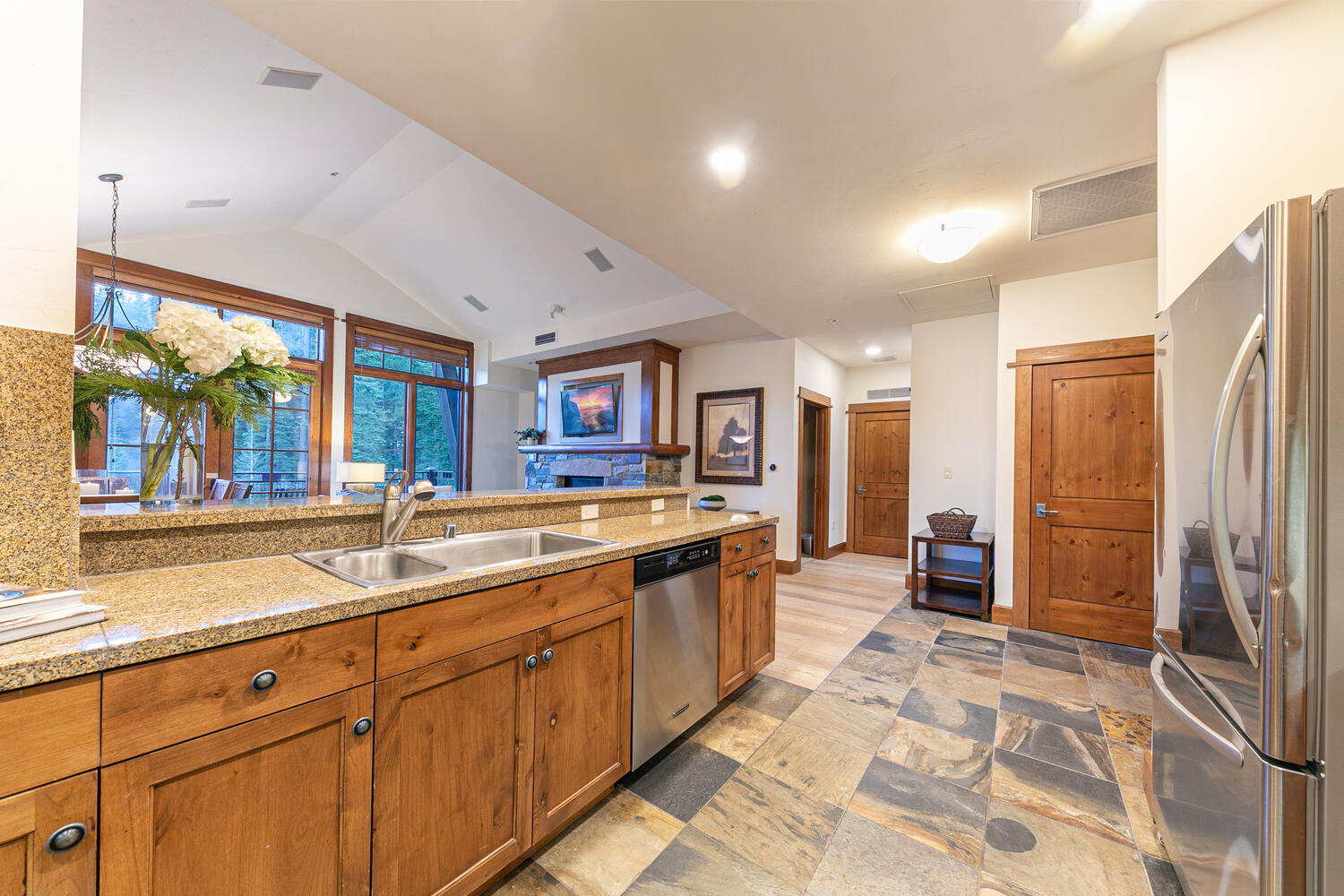 This kitchen features warm-toned wood cabinetry paired with speckled stone countertops and a stainless steel sink. The space is anchored by a slate-style tile floor and opens into a living area with a stone-clad fireplace and large windows that provide a view of the outdoors. The perspective captures the functional kitchen workspace in the foreground, transitioning into the open-concept living area beyond the breakfast bar.