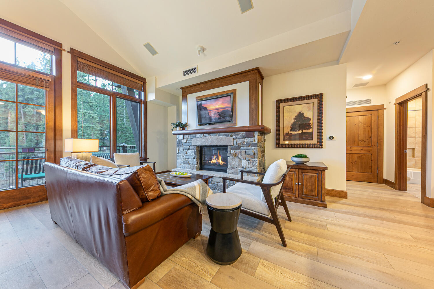This inviting living room features a warm, rustic aesthetic with light wood flooring and a prominent stone-clad chimney breast housing a fireplace and television. A plush brown leather sofa and a modern armchair are arranged around a central coffee table, creating a cozy seating area. Large windows and a glass door provide ample natural light and views of the surrounding forest, enhancing the serene atmosphere of the space.