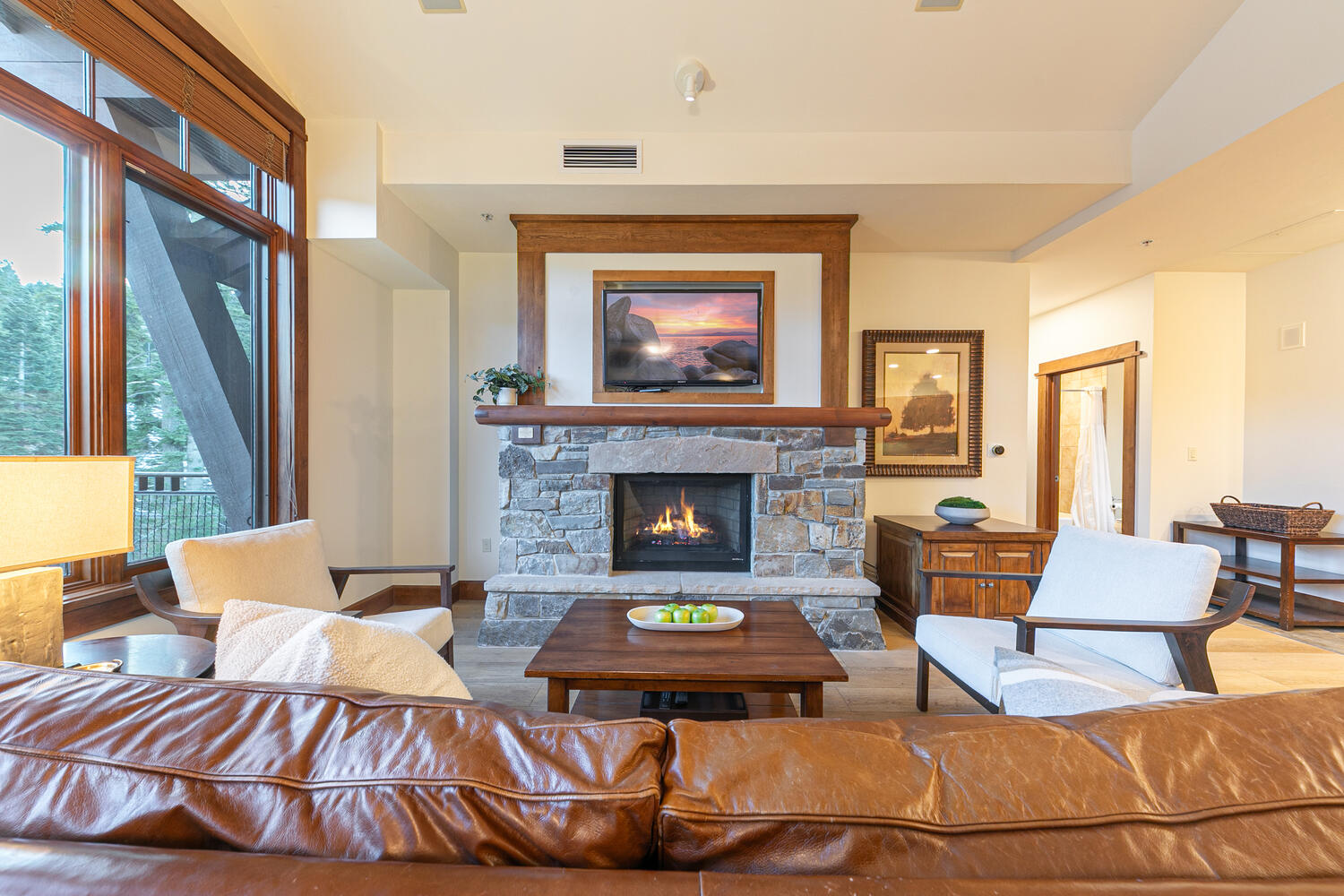 This inviting living room features a prominent stone-clad chimney breast with a built-in television and a cozy fireplace. The space is furnished with a large brown leather sofa in the foreground, two light-colored armchairs, and a central wooden coffee table topped with a bowl of green apples. Large windows to the left provide ample natural light and a view of the outdoors, creating a warm and comfortable atmosphere.