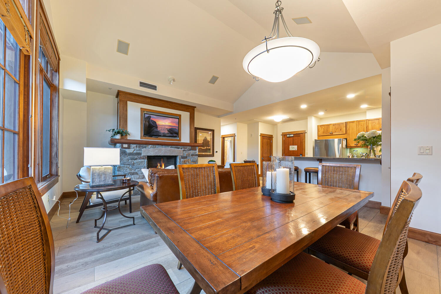 This open-concept living and dining area features a warm, rustic aesthetic with a large wooden dining table as the central focus. Beyond the dining space, a stone-clad chimney breast with a built-in television creates a cozy focal point for the adjacent living area, which includes a leather sofa and a side table with a lamp. The room is illuminated by a prominent modern pendant light fixture and recessed lighting, with light-toned wood flooring throughout and a view into the kitchen area in the background.
