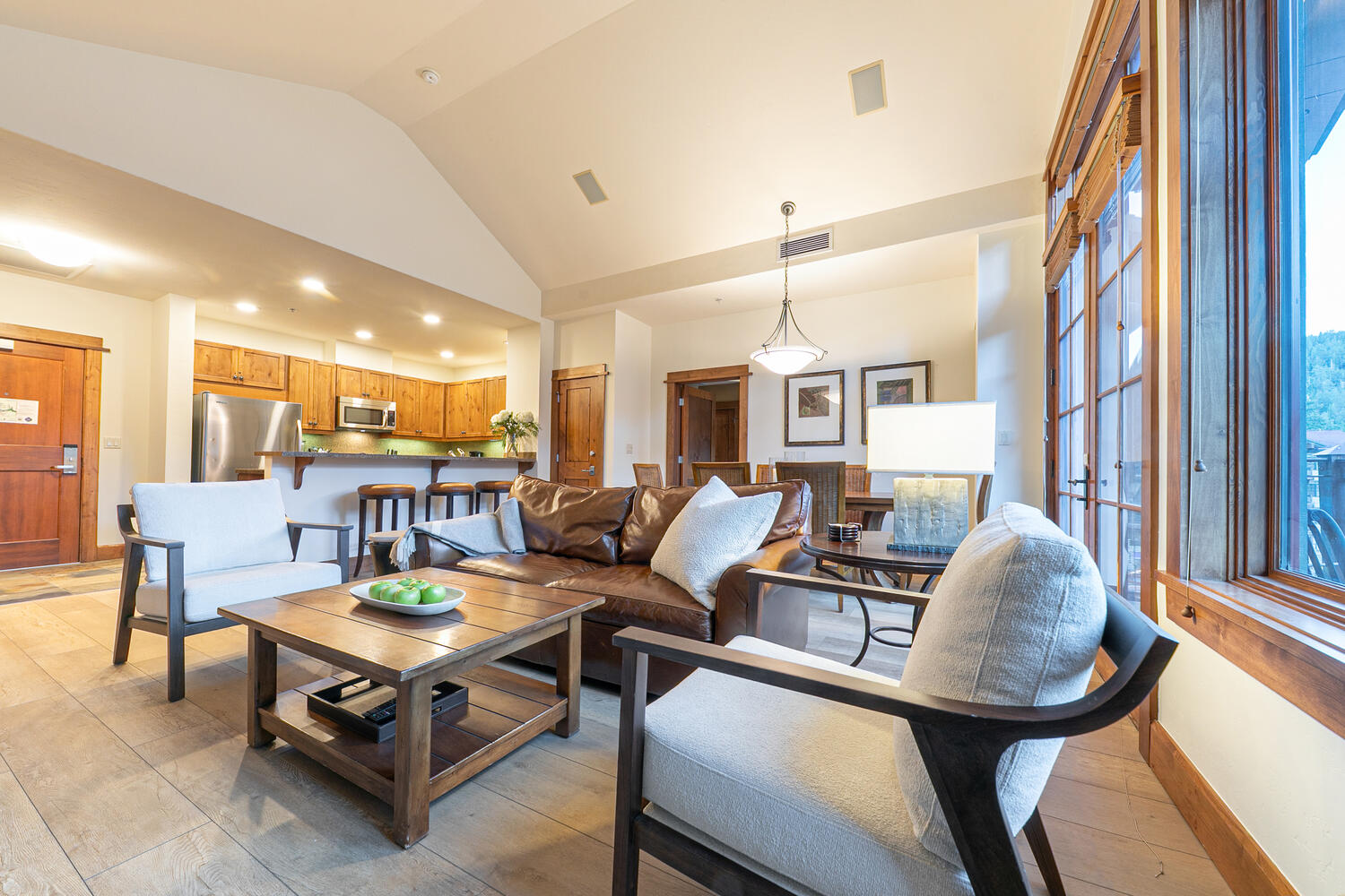 This open-concept living area features a vaulted ceiling and a warm, rustic aesthetic with light wood flooring and natural wood trim. The space is anchored by a large brown leather sofa, two light-colored armchairs, and a wooden coffee table, with a kitchen and breakfast bar visible in the background. Large windows along the right wall provide ample natural light, creating an inviting and spacious atmosphere suitable for entertaining.