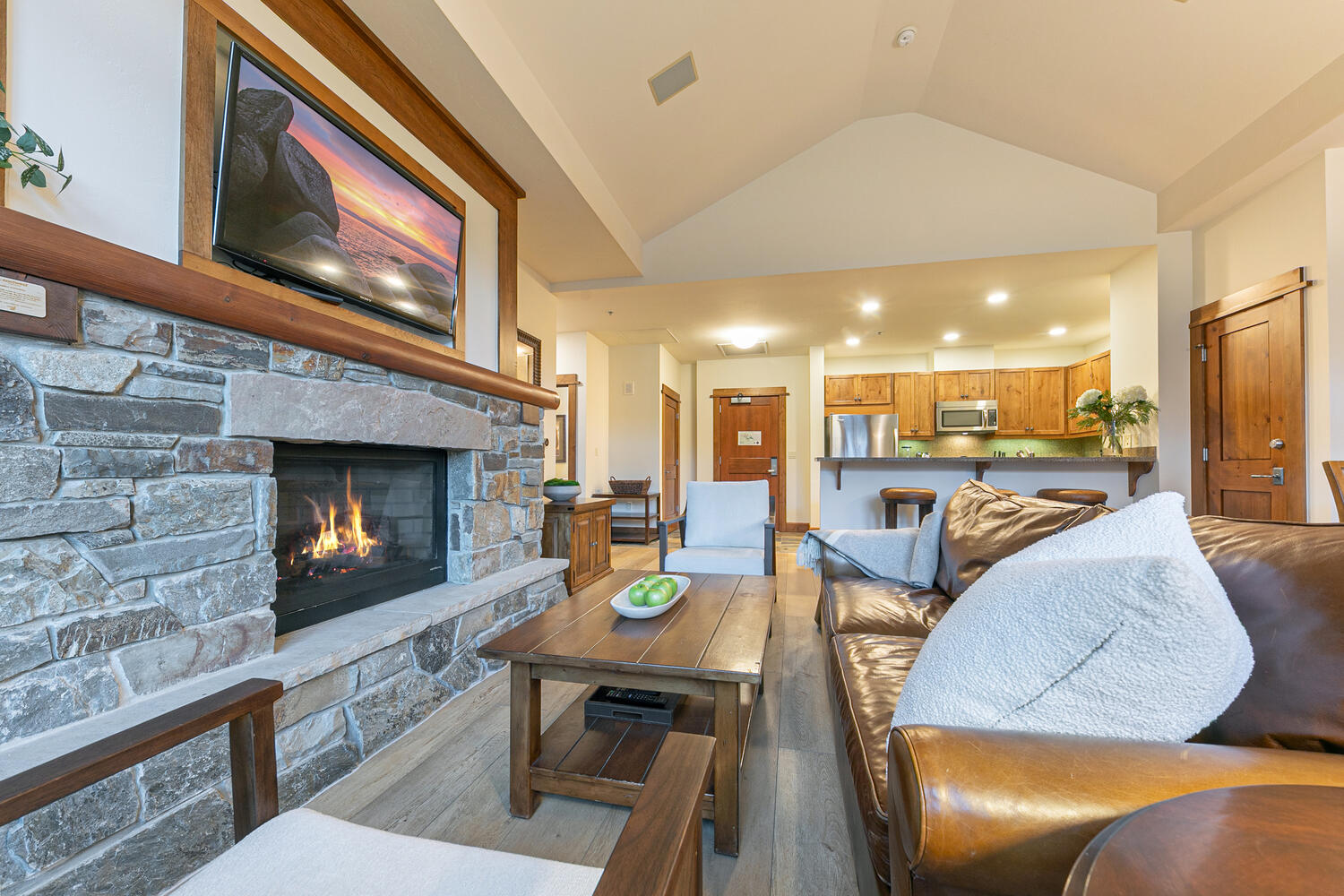This inviting living room features a prominent stone-clad chimney breast with a built-in fireplace and a mounted television above. The space is anchored by a large leather sofa with plush white cushions and a wooden coffee table, creating a warm, rustic atmosphere. The open-concept layout flows seamlessly into a kitchen area in the background, highlighting the home's spacious and connected design.
