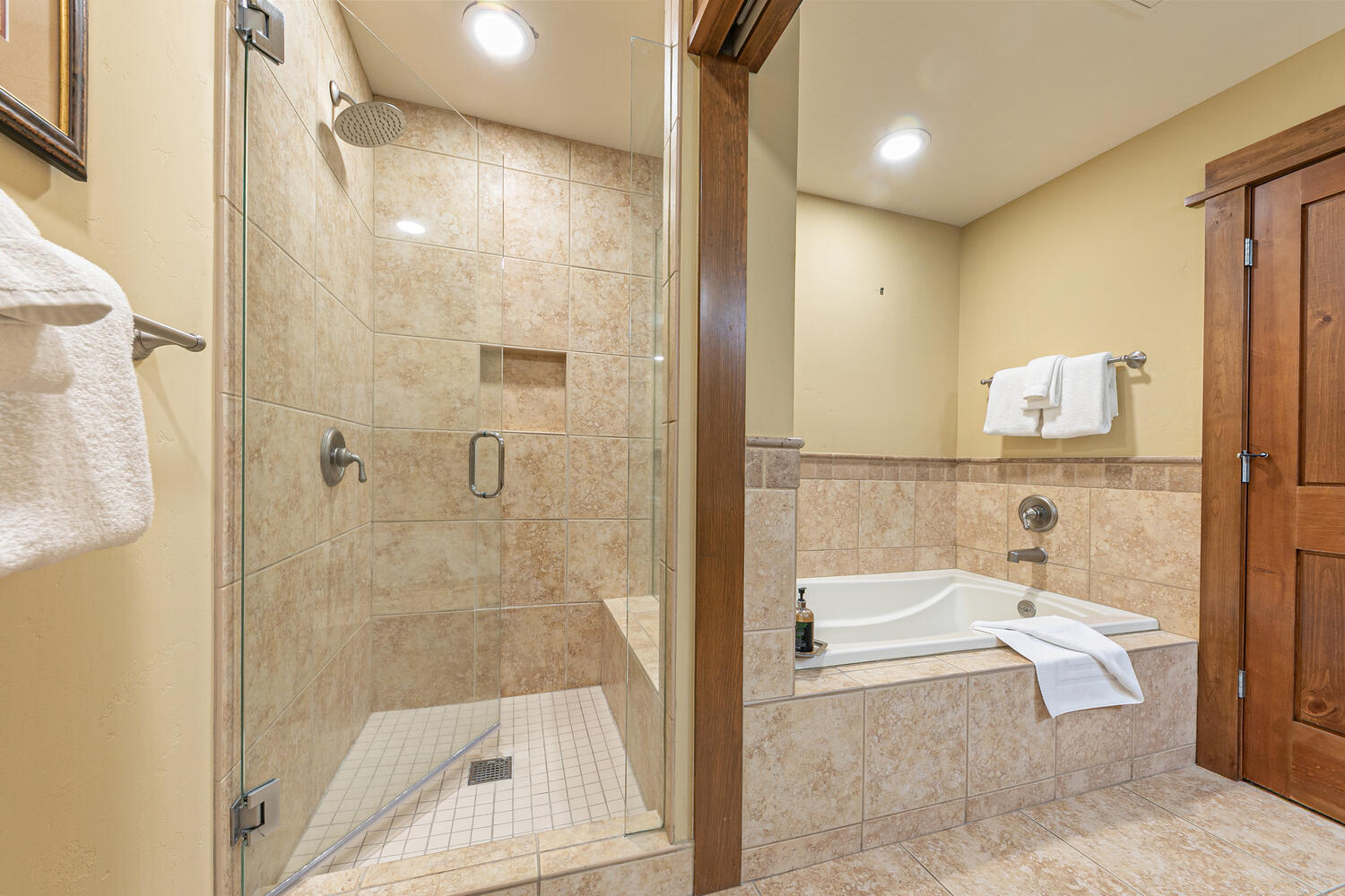 This bathroom features a glass-enclosed walk-in shower with a built-in bench and a separate soaking tub, both finished in neutral-toned stone tile. The space is accented by warm-toned walls, a wooden door, and recessed lighting, creating a cohesive and inviting atmosphere. The perspective captures both bathing areas, highlighting the functional layout and clean, classic design.