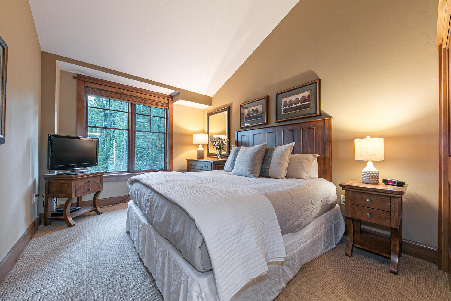 This cozy bedroom features a queen-sized bed with a dark wood headboard, neutral bedding, and a white ruffled bed skirt. The room is accented by warm-toned walls, a wooden nightstand with a lamp, and a matching wooden media console holding a television. Large windows provide a view of the surrounding forest, creating a peaceful and inviting atmosphere.