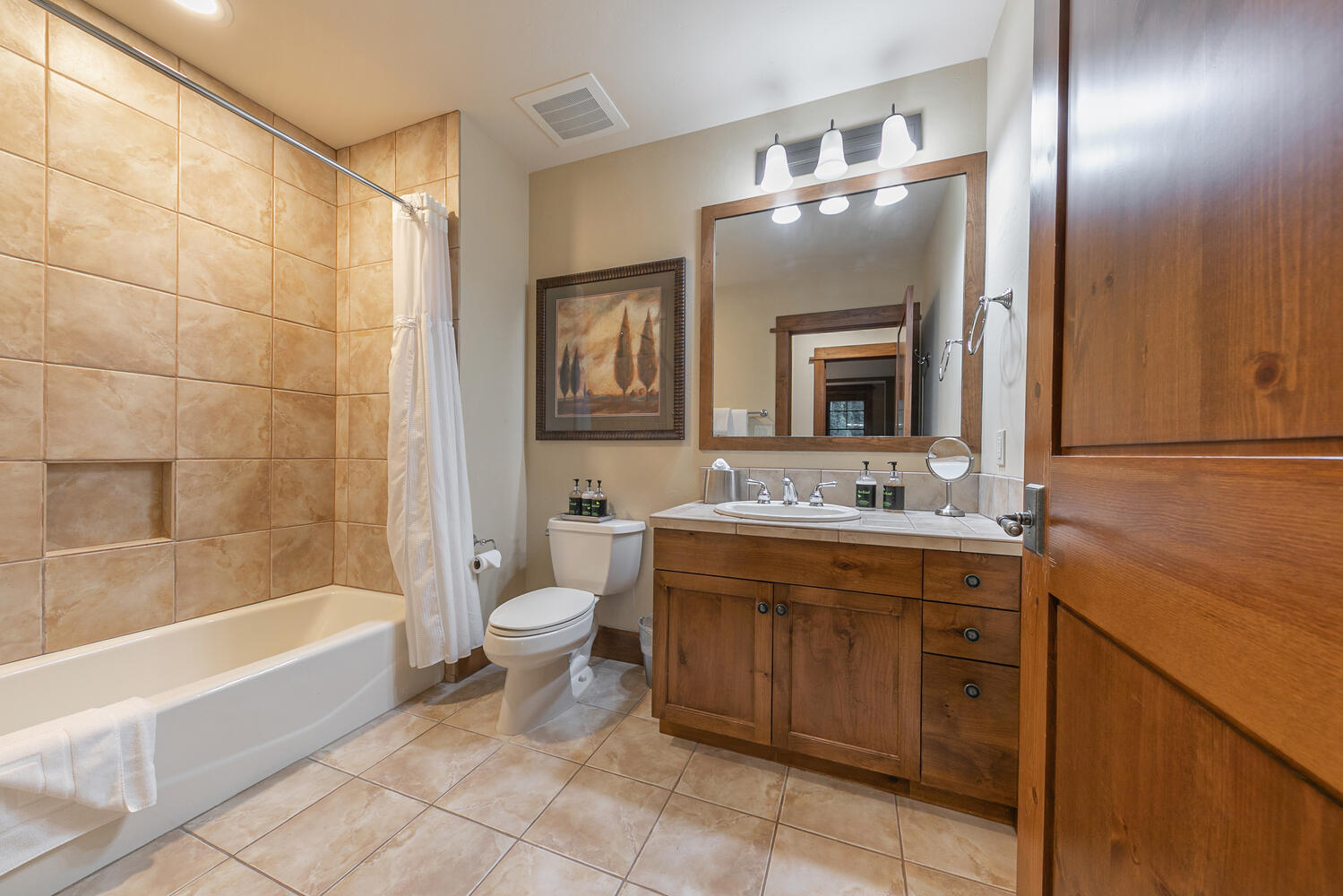 This bathroom features a warm, rustic aesthetic with light-toned tile flooring and matching wall tiles in the shower-tub combination. The space includes a wooden vanity with a stone countertop, a framed mirror, and a toilet positioned between the vanity and the tub. The perspective is a wide-angle shot from the doorway, capturing the functional layout and cozy, neutral color palette.