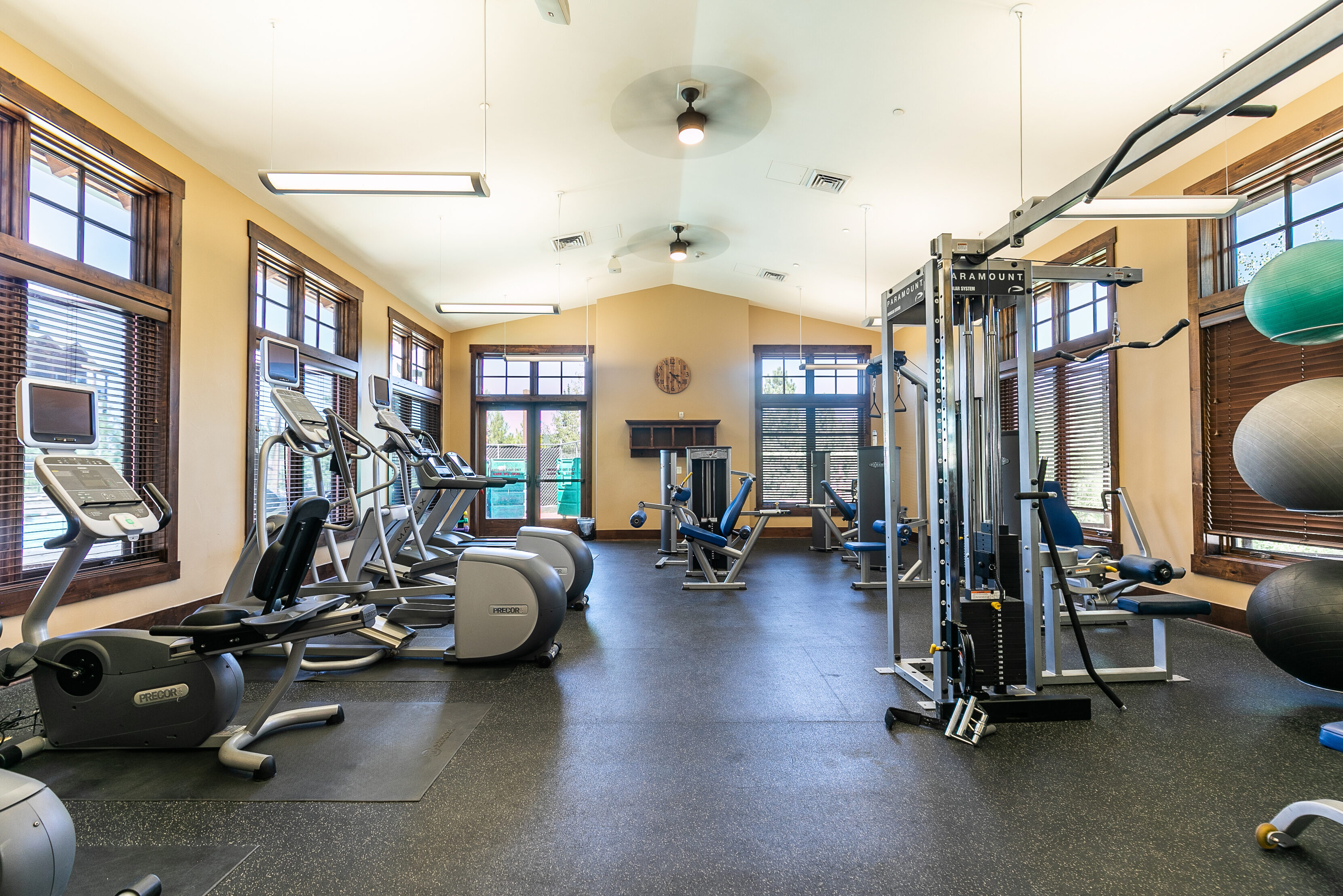 This well-equipped home gym features a variety of professional-grade exercise machines, including elliptical trainers and a multi-station weight system. The room is brightened by large windows with wooden blinds that allow natural light to fill the space, while the dark, durable flooring provides a functional surface for workouts. A vaulted ceiling with dual fans and a central wall clock complete the clean, organized aesthetic of this fitness area.