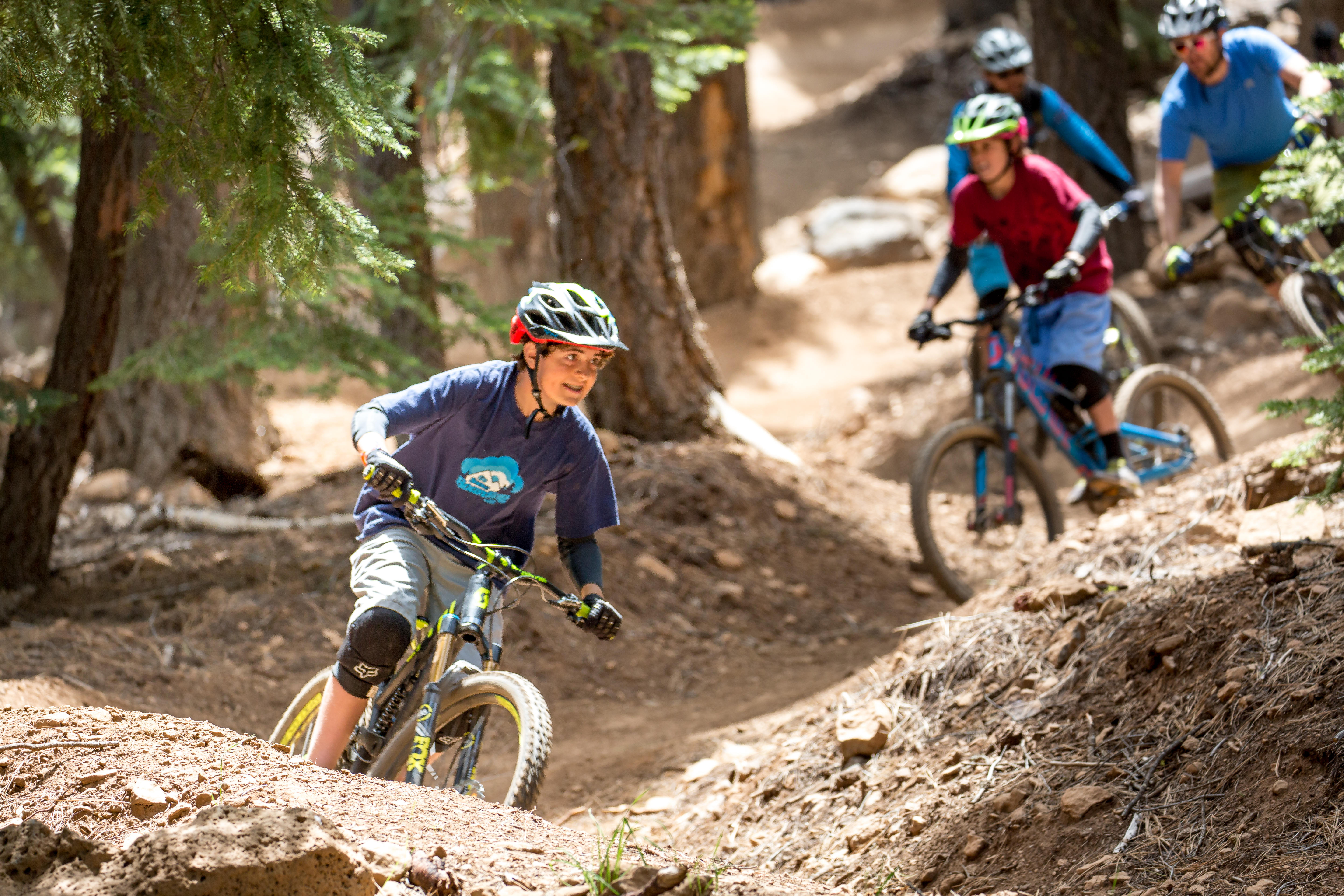This action-oriented image captures a group of mountain bikers navigating a dusty, winding dirt trail through a dense forest. The perspective is low and dynamic, focusing on the lead rider in the foreground while others follow along the path behind. The setting highlights a rugged, natural outdoor recreational area, emphasizing the terrain and active lifestyle potential of the location.