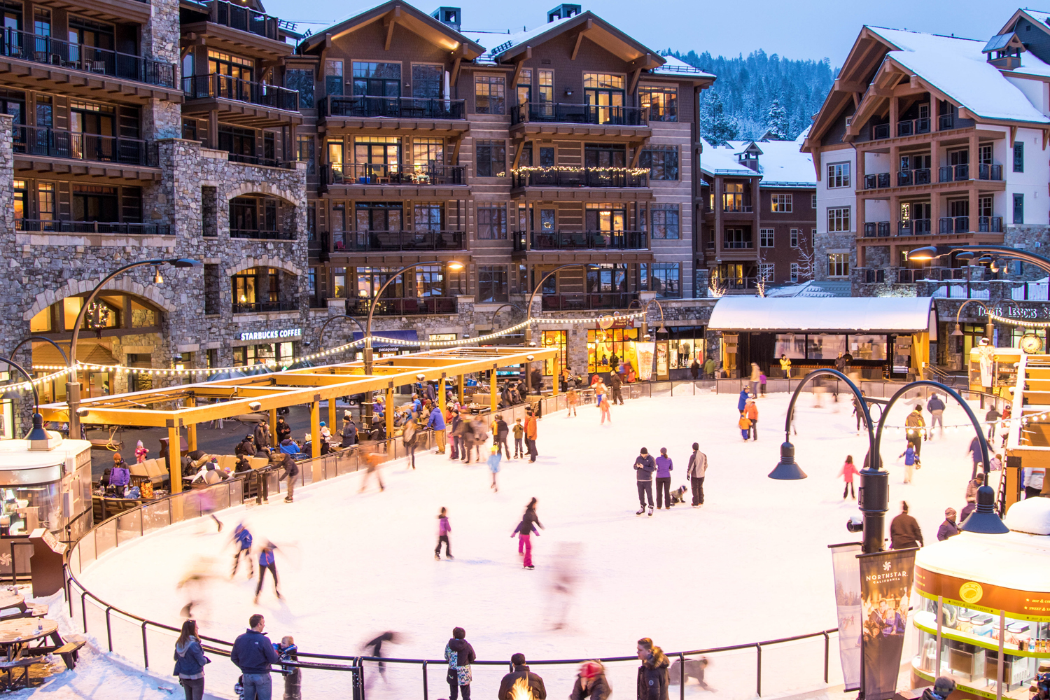 This vibrant scene captures a bustling outdoor ice skating rink nestled within a luxury mountain resort village. The architecture features multi-story buildings with stone facades, timber accents, and numerous balconies, creating a cozy, upscale alpine atmosphere. The perspective is elevated, looking down over the rink where people are skating, while warm lighting from the surrounding shops and string lights adds a welcoming, cinematic glow to the evening setting.