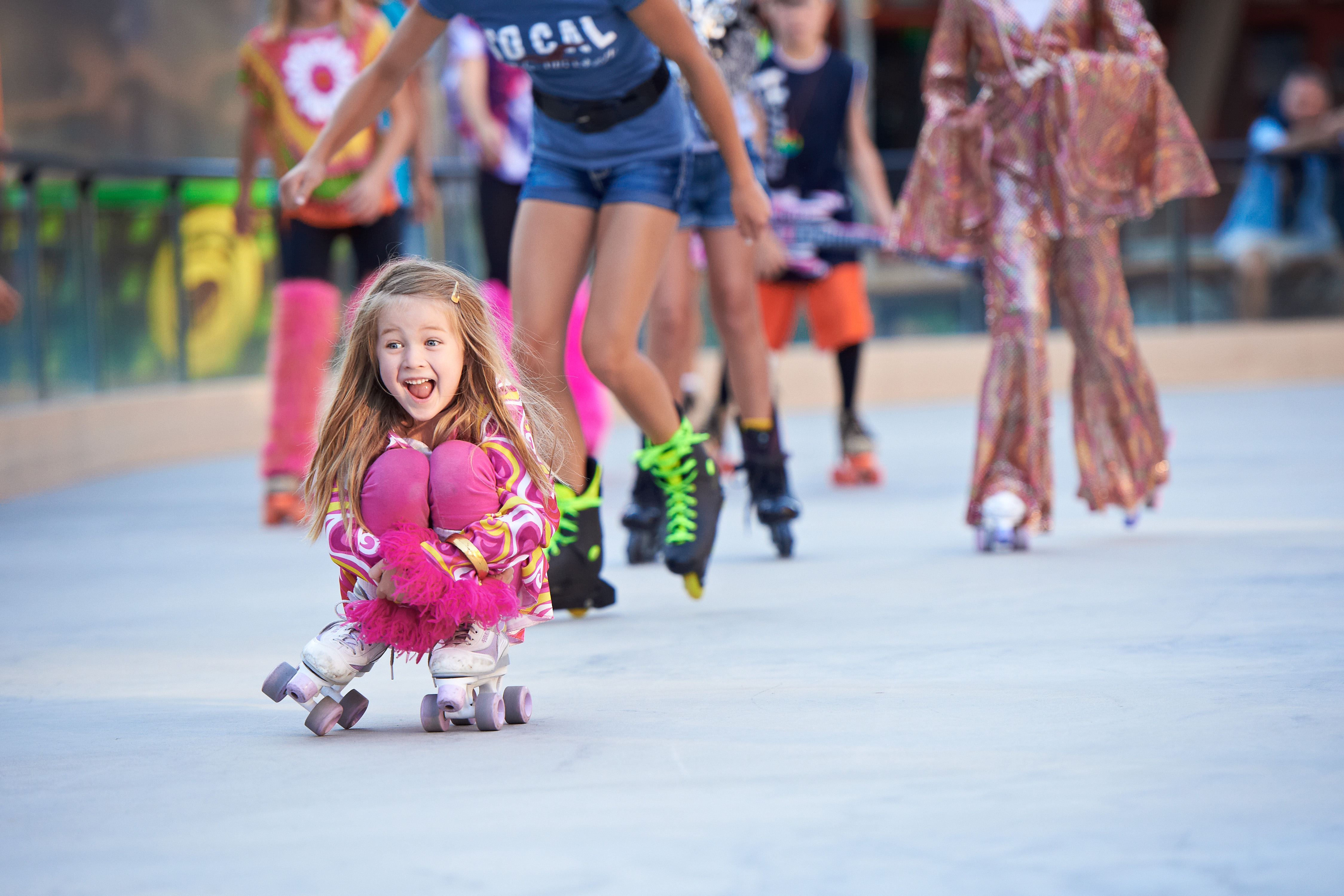 This image captures a vibrant, outdoor roller skating rink, likely part of a community recreational facility. A young girl in a colorful patterned outfit is in the foreground, joyfully skating in a crouched position, while other skaters are visible in the background on the smooth, paved surface. The scene conveys an active, family-friendly atmosphere, highlighting the recreational amenities available to residents.
