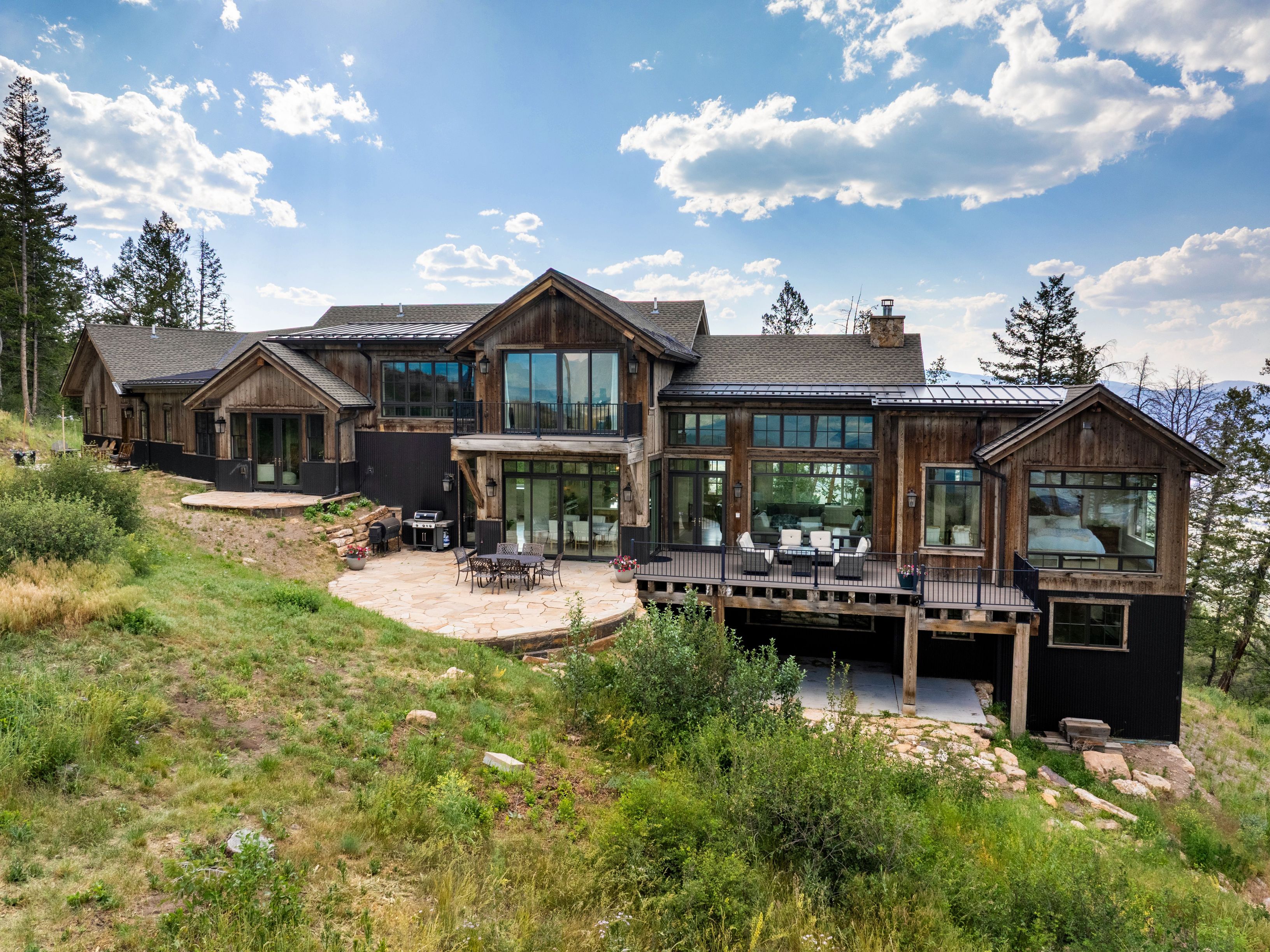 This stunning custom rustic-modern home features a striking combination of weathered timber siding and dark metal accents. The expansive exterior showcases a large flagstone patio for outdoor entertaining, a spacious elevated deck with sleek iron railings, and extensive glass windows that offer panoramic views of the surrounding landscape. The architectural design seamlessly blends a mountain lodge aesthetic with contemporary luxury, perfectly situated within a rugged, natural hillside environment.