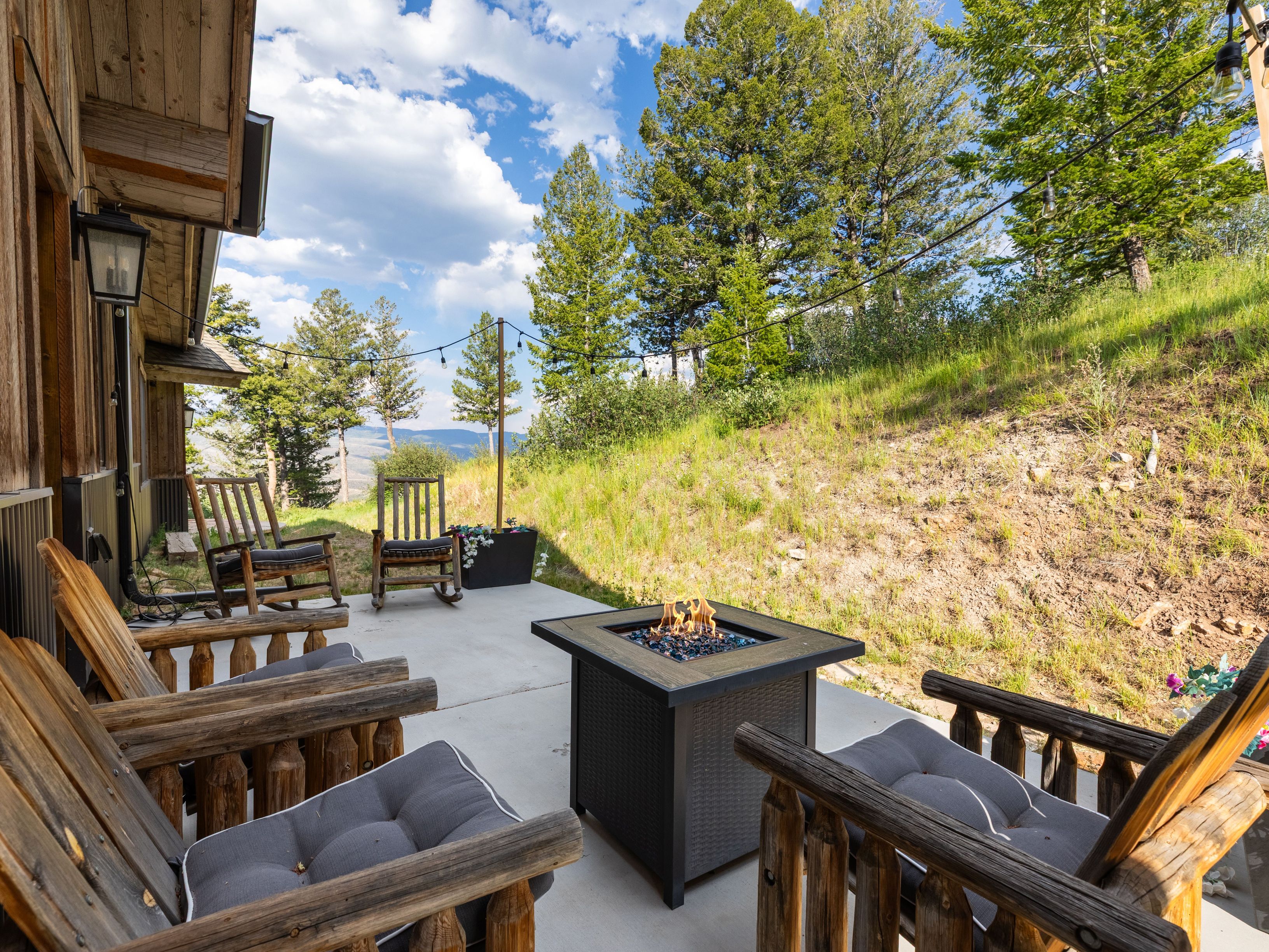 This cozy outdoor patio features a rustic mountain aesthetic with handcrafted log rocking chairs and a contemporary square fire pit. The concrete space is framed by string lights and overlooks a scenic, sloped landscape with trees, providing a relaxing area for evening gatherings. The attached wooden structure reflects a charming, cabin-inspired architecture that blends seamlessly with the natural surroundings.