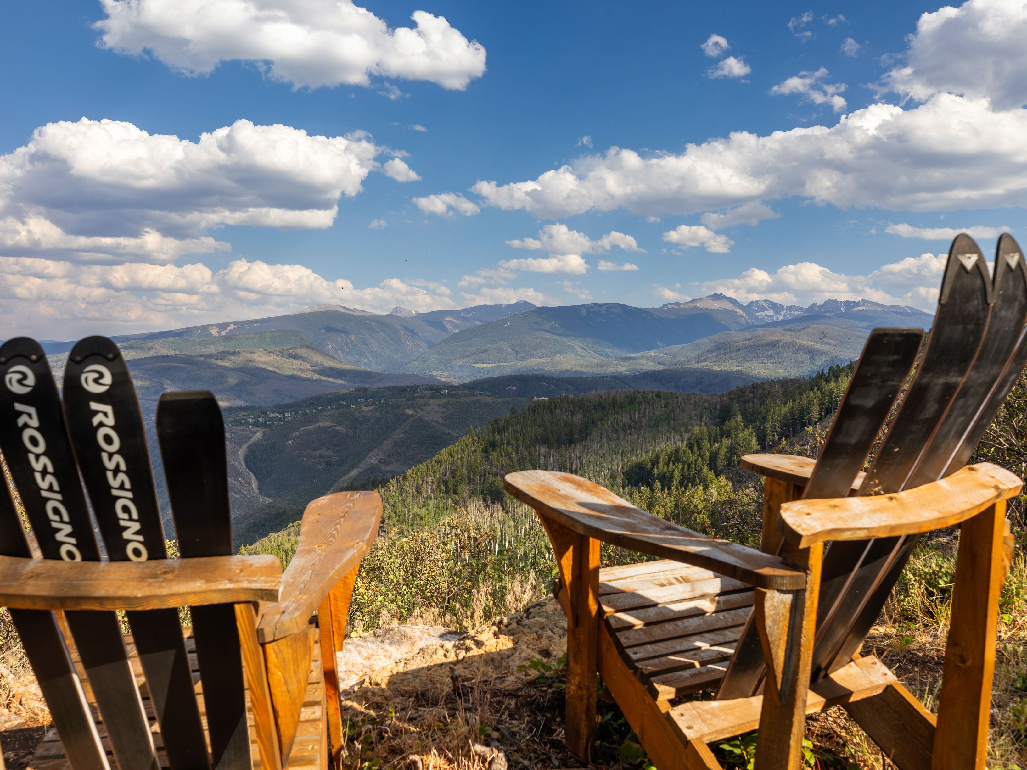 This scenic overlook features two rustic Adirondack chairs crafted with repurposed Rossignol skis as backrests, positioned on a natural rocky cliffside. The vantage point offers a breathtaking, unobstructed panoramic view of a vast mountain valley, dense forests, and distant snow-capped peaks under a bright, cloud-filled blue sky. It provides an ideal, tranquil setting for outdoor relaxation and highlights the scenic beauty associated with luxury mountain resort living.