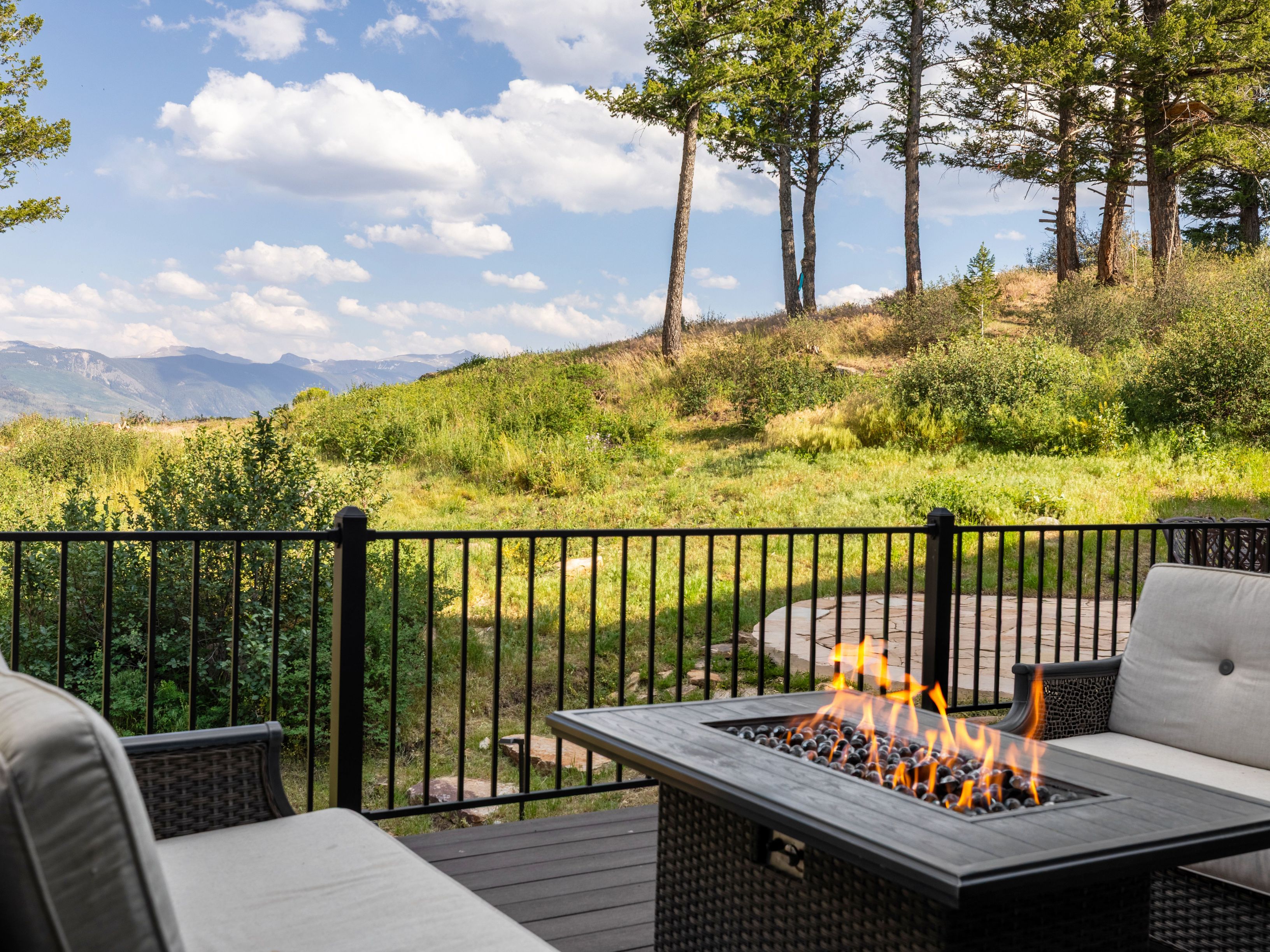 This inviting outdoor space features a dark wicker seating arrangement with light-colored cushions and a central rectangular fire table, perfect for cozy gatherings. The deck is enclosed by a black metal railing and offers breathtaking, unobstructed views of a lush mountain landscape and clear blue sky. It provides a serene and scenic setting for relaxation, highlighting the property's prime location for enjoying the natural environment.