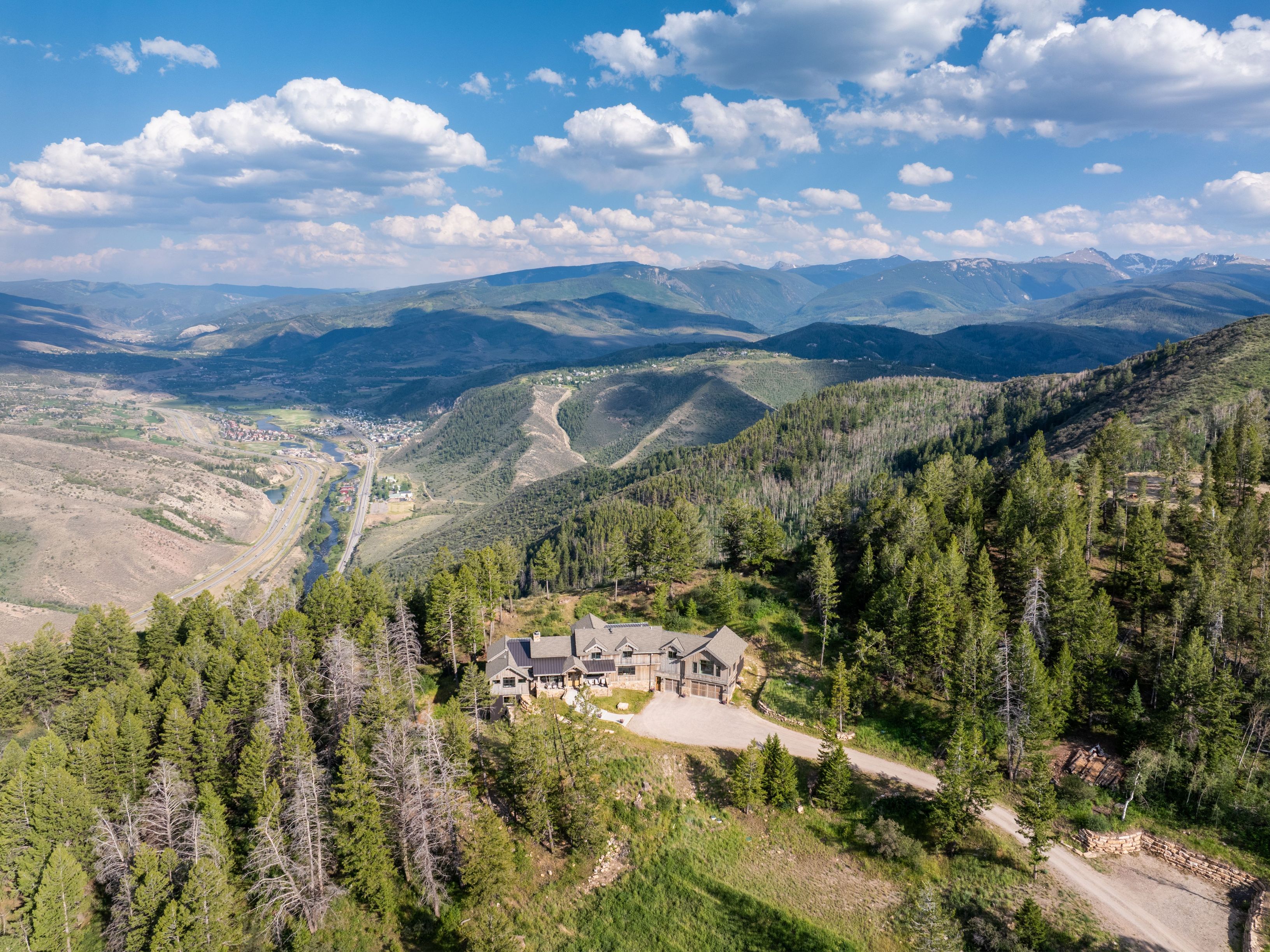 This aerial view captures a luxury mountain retreat nestled amidst a vast, forested landscape with sweeping valley and distant mountain range views. The residence features a classic, rustic architectural style with a multi-gabled roofline and a spacious gravel driveway. Its elevated position highlights the property's secluded atmosphere and stunning panoramic natural surroundings.
