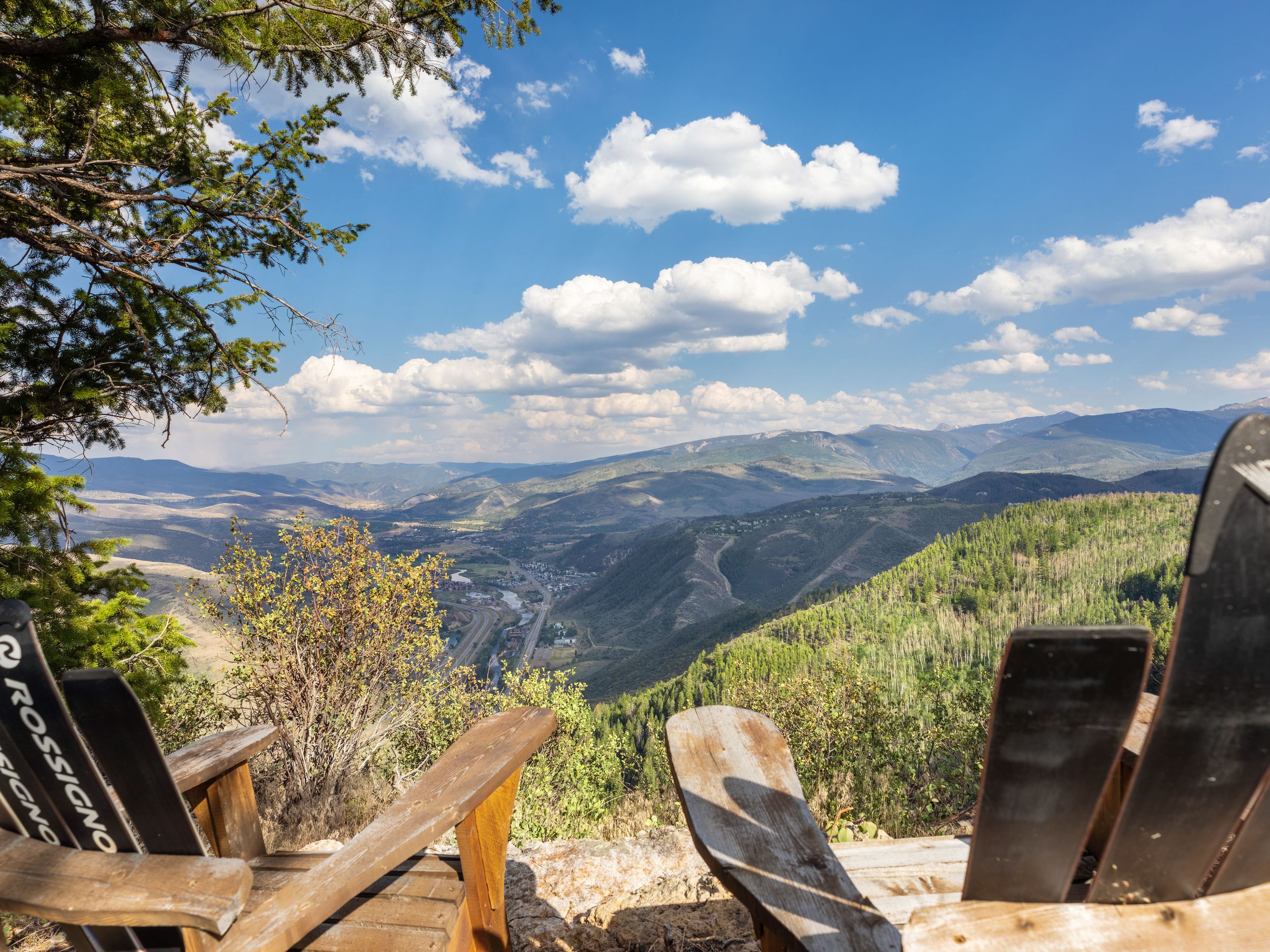 This image showcases a scenic outdoor overlook featuring unique, custom-made Adirondack-style chairs crafted from vintage Rossignol skis. The vantage point offers a breathtaking, panoramic view of a valley, winding roads, and distant mountain ranges under a bright blue sky. It serves as a compelling lifestyle shot that highlights a premium, high-altitude location with exceptional natural beauty.
