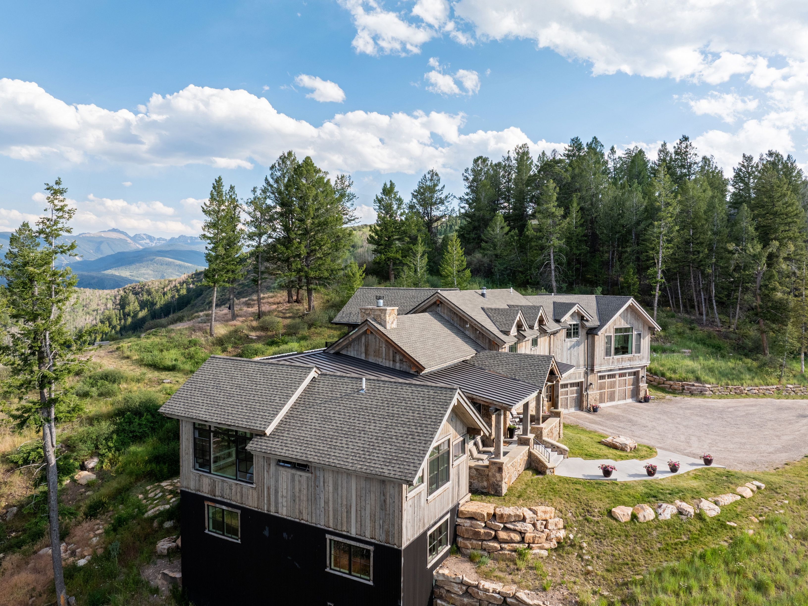 This elevated aerial perspective showcases a stunning multi-level mountain home designed with a rustic, modern aesthetic, featuring natural wood siding and stone accents. The residence is situated on a lush, hillside lot surrounded by dense forest, offering breathtaking panoramic views of the distant mountain ranges. The property includes a large gravel driveway, a spacious concrete patio, and intricate rooflines that integrate seamlessly with the natural landscape.
