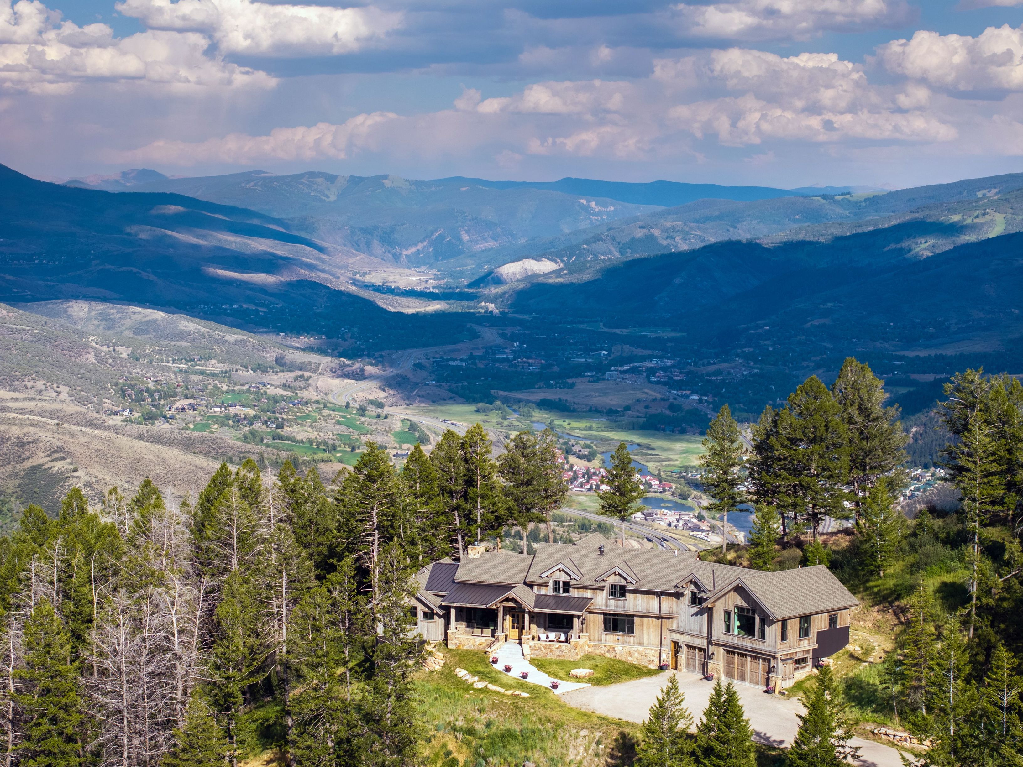 This stunning aerial perspective showcases a custom-built mountain home perched on a hillside, offering breathtaking panoramic views of the valley below. The residence features a rustic architectural style with wood siding, stone accents, and a complex gabled roofline, complemented by a spacious multi-car garage. Its elevated location provides unparalleled scenery, blending luxurious mountain living with a serene, natural environment.