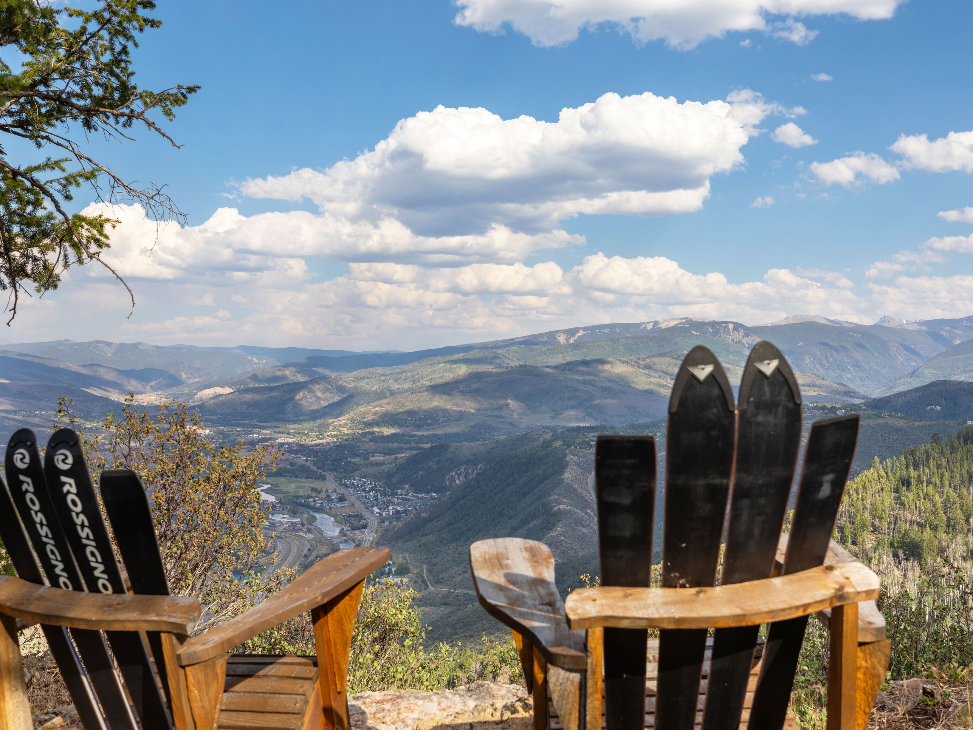 This image features a scenic mountain lookout point equipped with unique custom chairs constructed from repurposed Rossignol skis. These chairs are positioned to offer a stunning, elevated panoramic view of the valley, winding river, and distant mountain ranges below. This amenity highlights the outdoor lifestyle and recreational appeal of the surrounding community.
