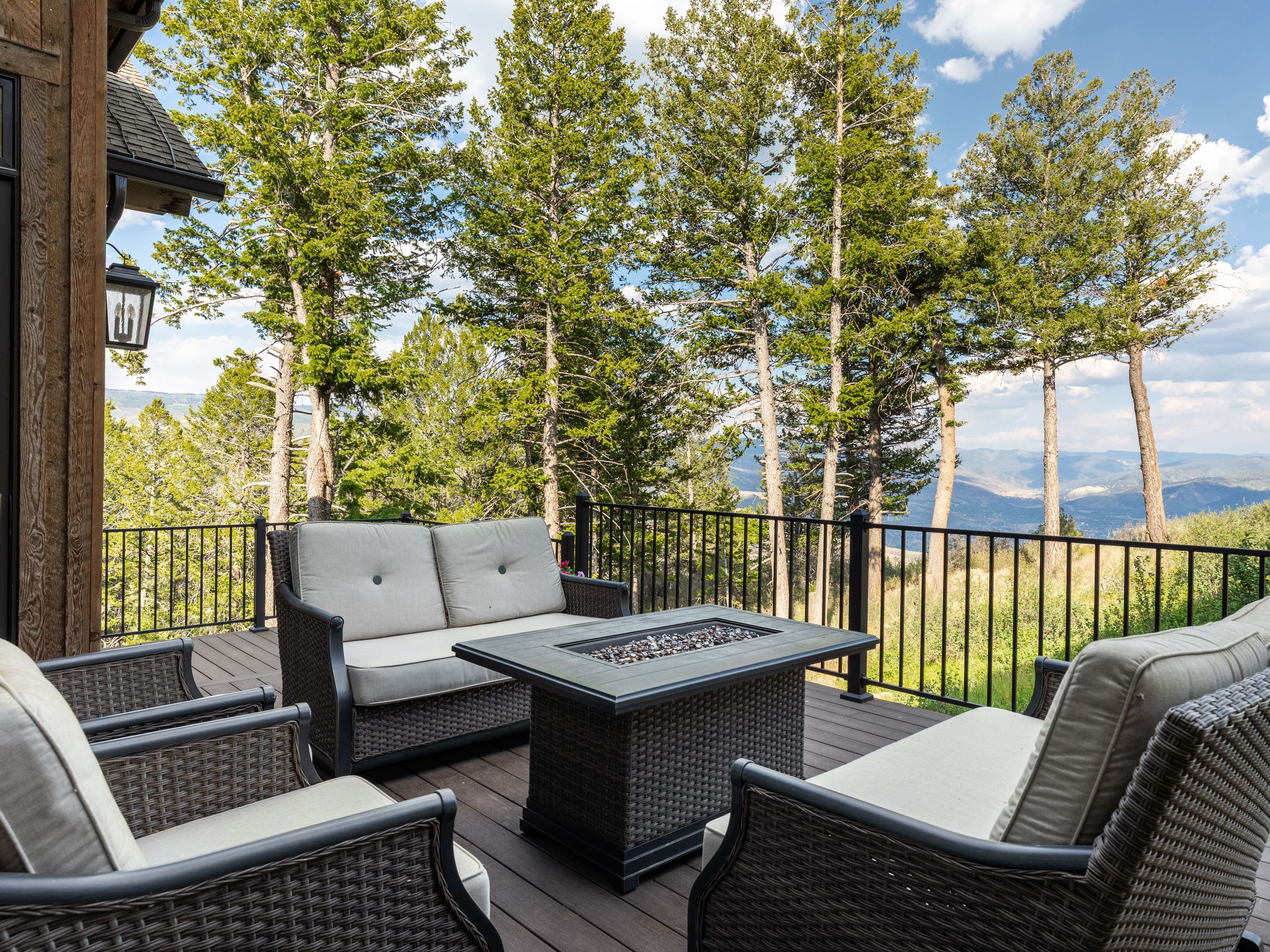 This inviting outdoor deck offers a serene mountain retreat with a cozy wicker furniture set, including a sofa and armchairs centered around a fire pit table. The space is defined by its dark wood decking and sleek black metal railing, which frames breathtaking, unobstructed panoramic views of the forested landscape and distant mountains. It provides a perfect, relaxed setting for outdoor entertaining or enjoying quiet moments amidst nature.