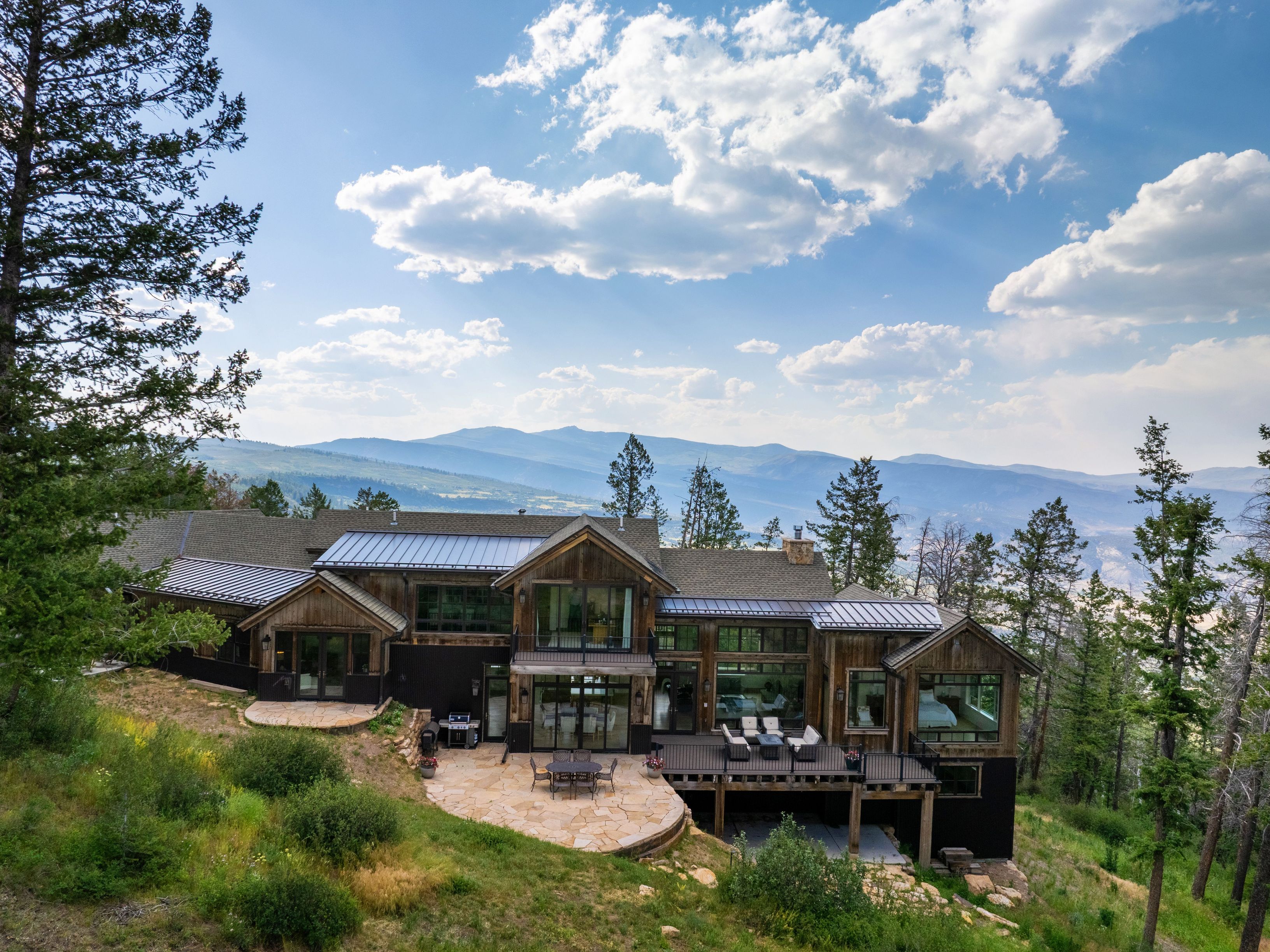This stunning custom mountain residence features a blend of natural wood siding and stone elements, perfectly integrated into its sloping landscape. The rear exterior offers expansive views of the valley and mountains, complemented by a stone patio, an elevated deck, and large floor-to-ceiling windows that invite natural light. The architectural style captures a rustic yet luxurious aesthetic, ideal for high-end mountain living.