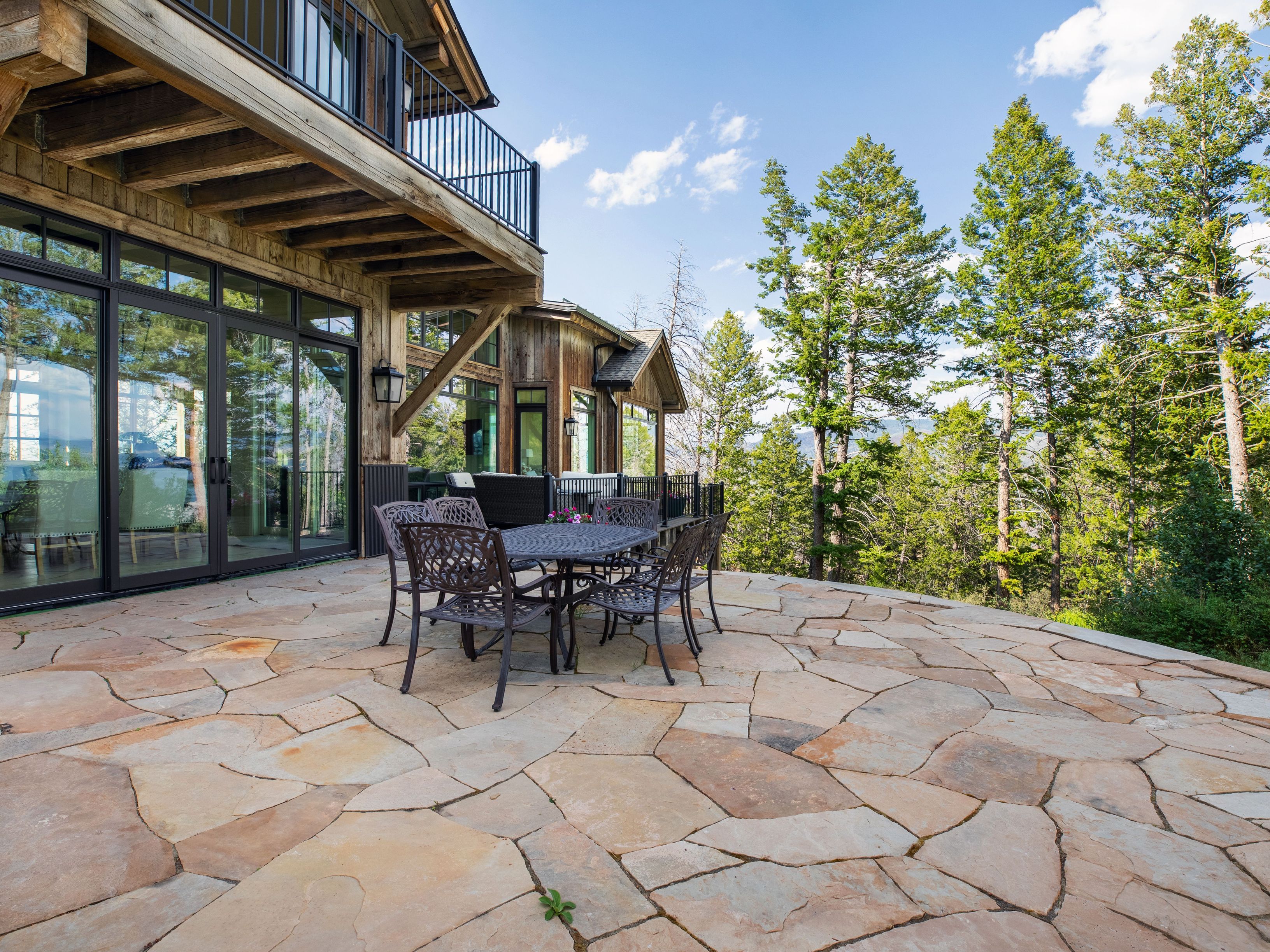 This expansive stone patio features a rustic outdoor dining set, providing a perfect vantage point for the surrounding forest and mountain views. The home showcases a rugged, wood-sided exterior with large glass doors that seamlessly connect the indoor living space to the outdoor terrace. The scene is bathed in bright, natural daylight, emphasizing the peaceful and secluded atmosphere of this mountain retreat.