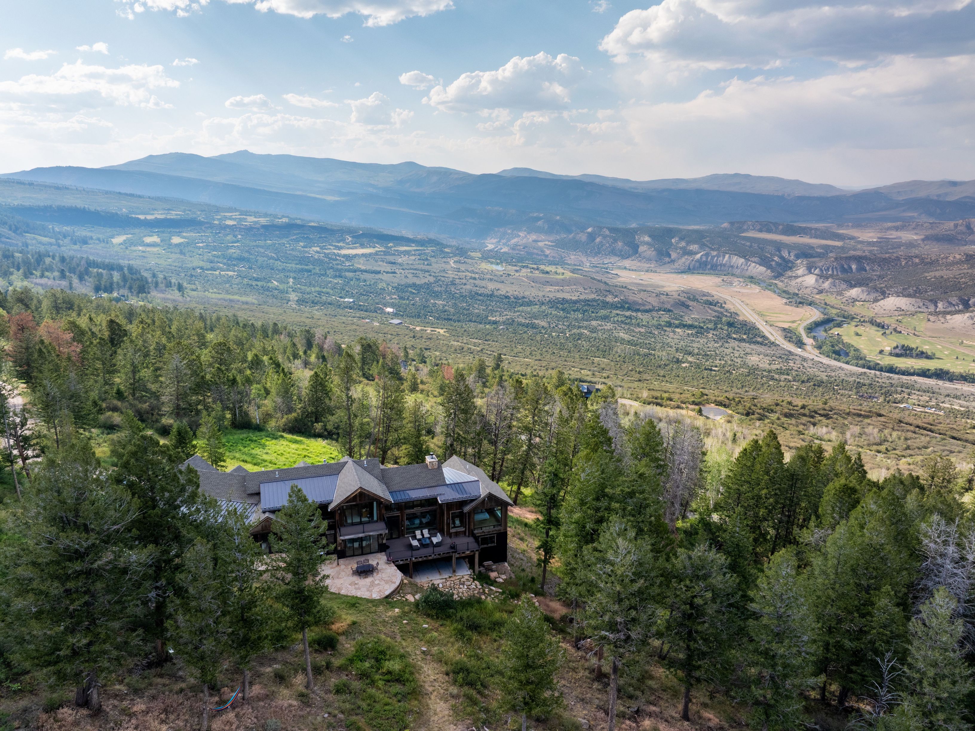 This stunning aerial view captures a luxury mountain retreat nestled amidst a dense forest on a hillside. The expansive multi-level home features a blend of natural wood and dark metal roofing, complemented by a large stone patio and a spacious elevated wooden deck. It offers breathtaking, panoramic vistas of the sweeping valley below, showcasing an ideal setting for serene, high-end mountain living.