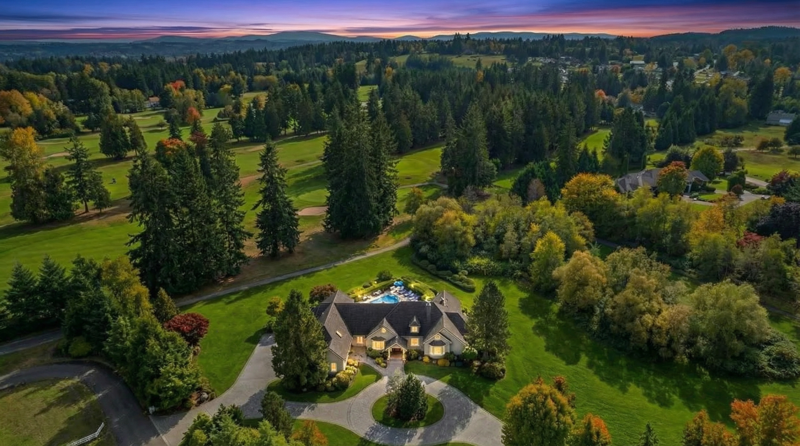 This is an aerial view of a luxury home nestled in a lush, green landscape, possibly a golf course community. The home features a well-maintained lawn, a circular driveway, and a swimming pool visible in the backyard. The setting suggests privacy and exclusivity, enhancing the property's appeal as a high-end real estate offering.