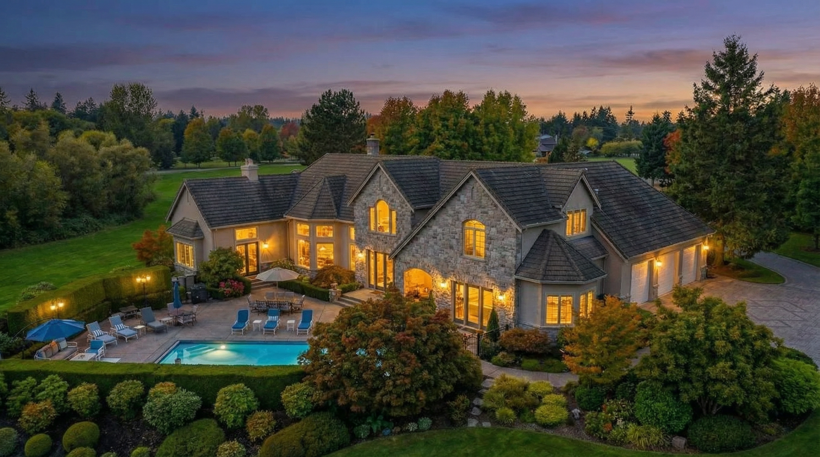 This image captures the front exterior of a grand two-story home at dusk. The property features a stone facade, a multi-car garage, and expansive landscaping with a swimming pool and patio area complete with lounge furniture. Warm lighting emanates from the windows creating an inviting ambiance and showcasing the architectural details of the residence.