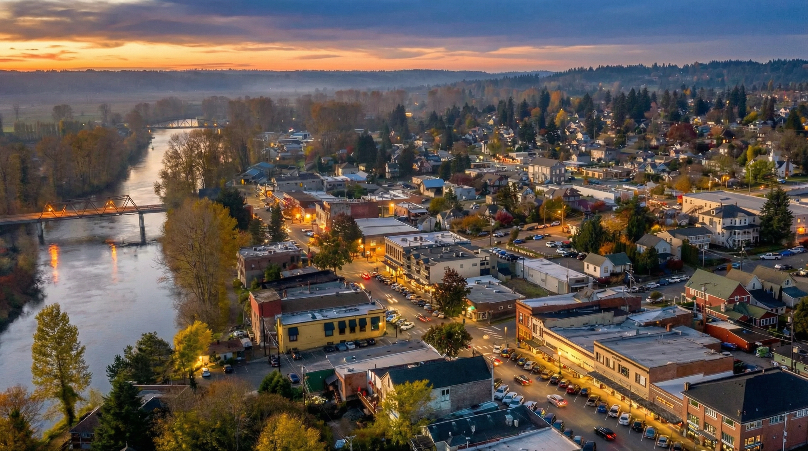 This aerial view showcases a charming town nestled beside a river, with two bridges adding character to the landscape. The town features a mix of commercial buildings, residences, and mature trees. The golden hour lighting enhances the aesthetic appeal, suggesting a peaceful and inviting community atmosphere that would likely attract potential buyers.