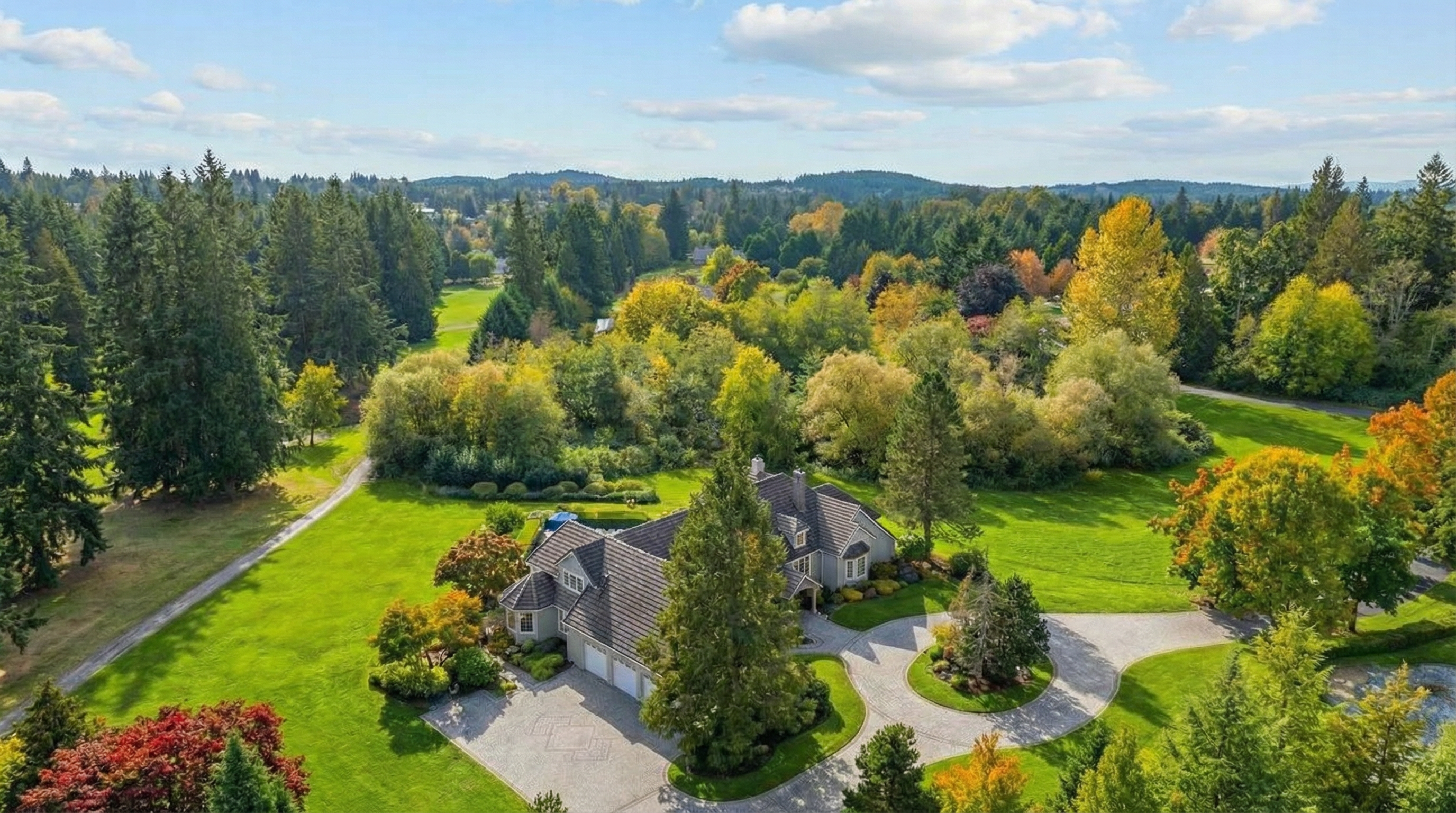 This is an aerial shot of a luxurious estate featuring a large house, a sprawling green lawn, and mature trees in autumn colors. The circular driveway adds to the property's grandeur. The view showcases a well-maintained landscape and a sense of privacy.