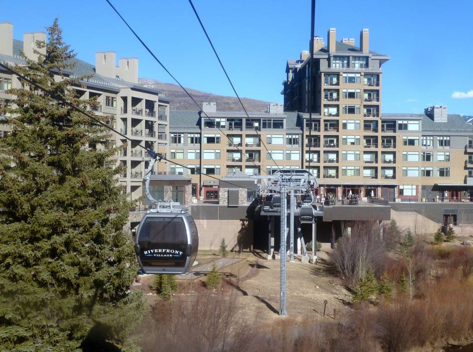 This image presents an exterior aerial view centered on a gondola transportation system serving a large, multi-story lodge or resort complex. The architecture features a blend of natural stone, wood elements, and large windows consistent with a mountain ski destination, with the facility seamlessly integrated into the landscape. This view highlights the convenient access to transit and the well-maintained outdoor surroundings of the Riverfront Village property.