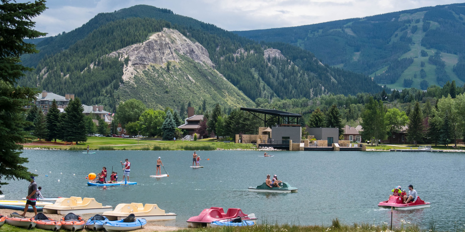 This scenic lakefront park serves as a prime community recreation area, featuring a spacious pond utilized for paddleboarding, kayaking, and pedal boating. In the background, a modern structure and lush forested mountains provide a picturesque, tranquil setting that highlights the recreational lifestyle of the community.