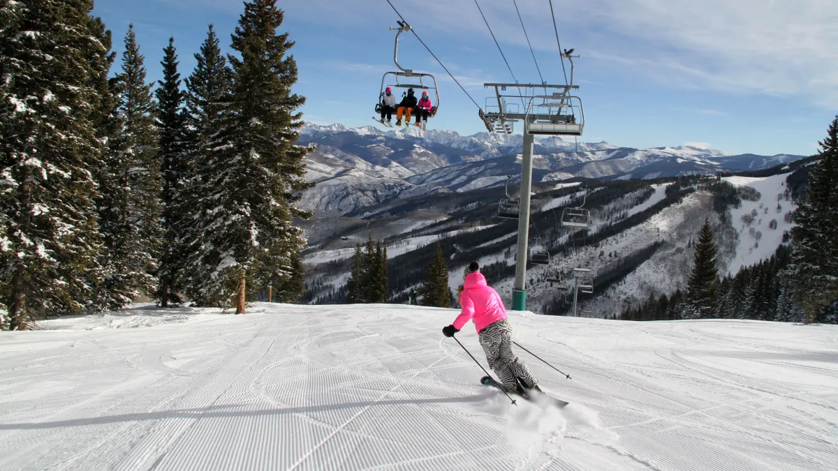 The image depicts a ski resort with a skier on a groomed slope, a ski lift in operation, and snow-covered mountains in the background. Tall evergreen trees line the side of the slope, adding to the scenic winter landscape. The scene highlights recreational opportunities and the access to ski facilities within the community.