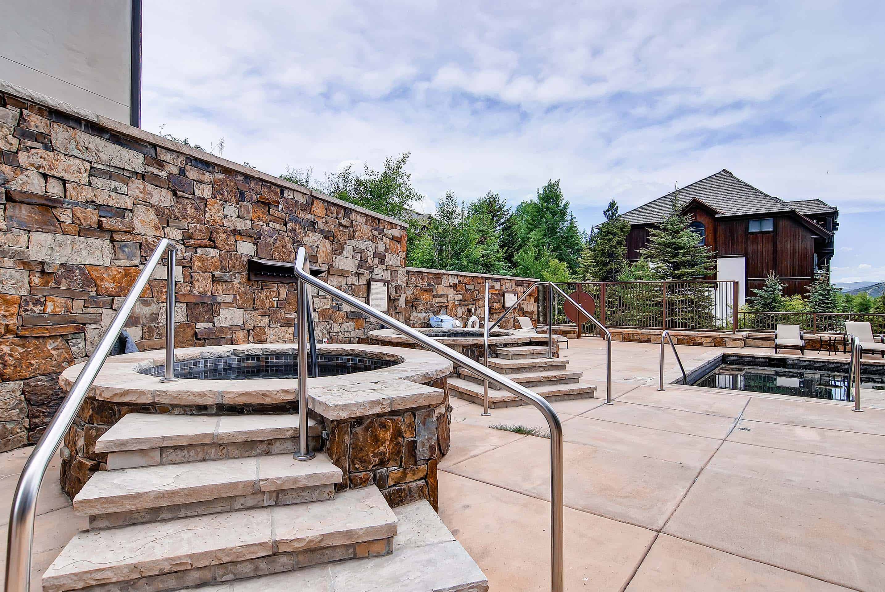 This image showcases a luxury pool and spa area. The space features multiple hot tubs with stone accents and stainless steel railings, along with a rectangular pool. The design incorporates natural stone elements and a patio area overlooking the surrounding landscape.