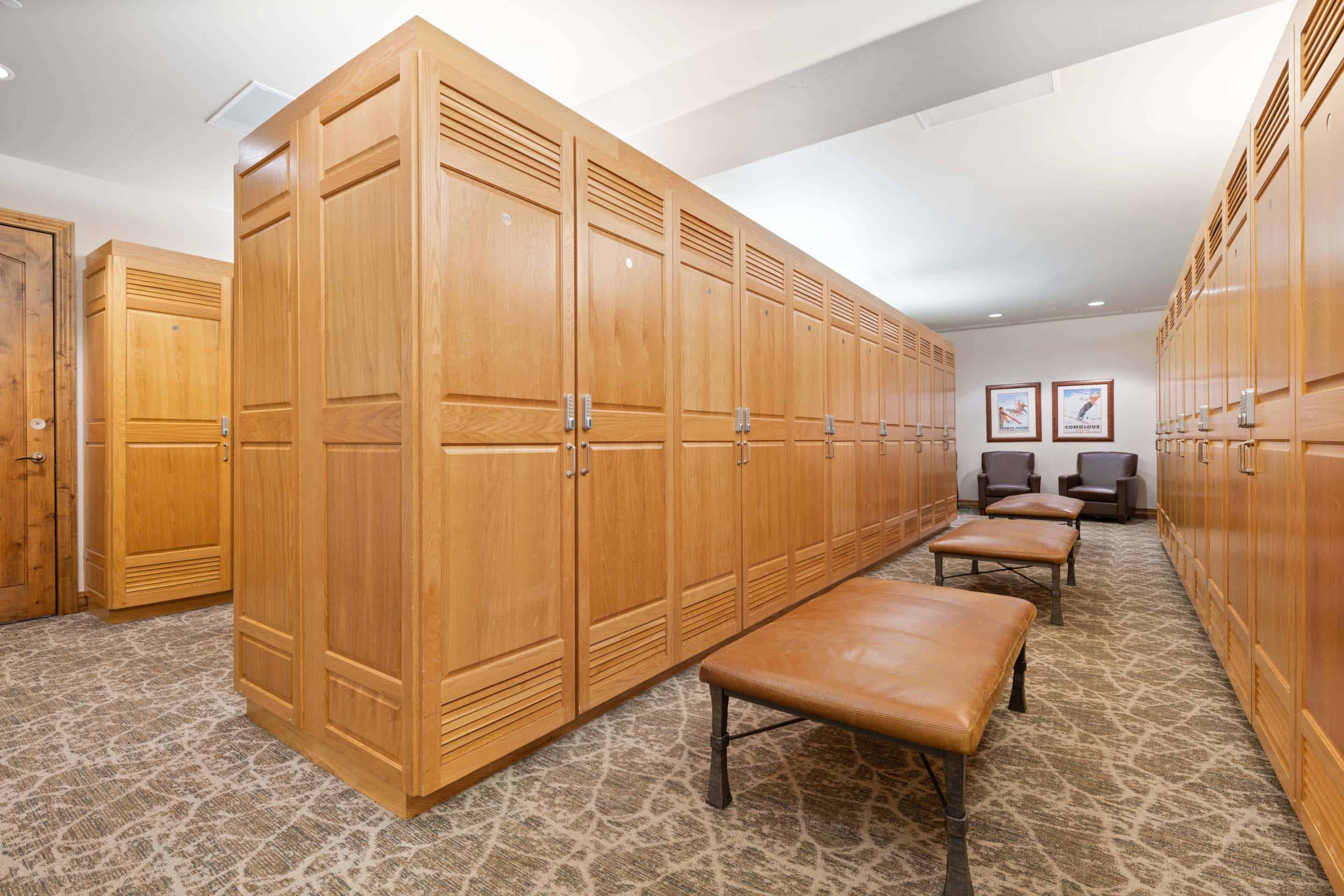 The image showcases an interior hallway featuring a long row of wooden lockers along the walls. Brown leather benches are available for seating in the middle of the room. Neutral-toned patterned carpet covers the floor, and framed artwork adorns the walls in the background, creating a functional yet aesthetically pleasing space.