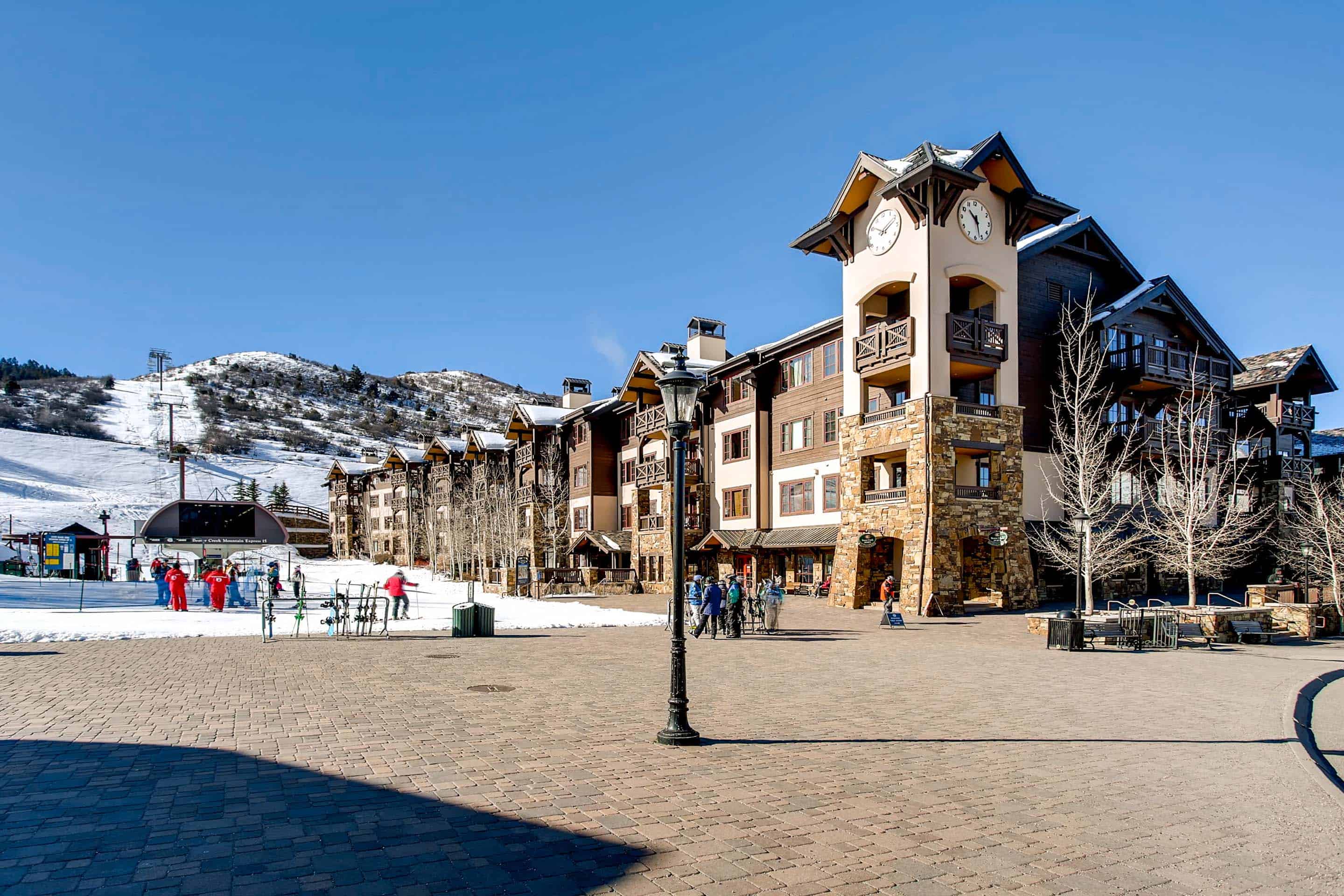 This is a front exterior shot of a multi-story resort or condominium building. The building features a stone and wood facade, multiple balconies, and a prominent clock tower. The scene is bustling with pedestrian activity and offers access to skiing facilities, suggesting a prime location and convenient access for residents or guests.