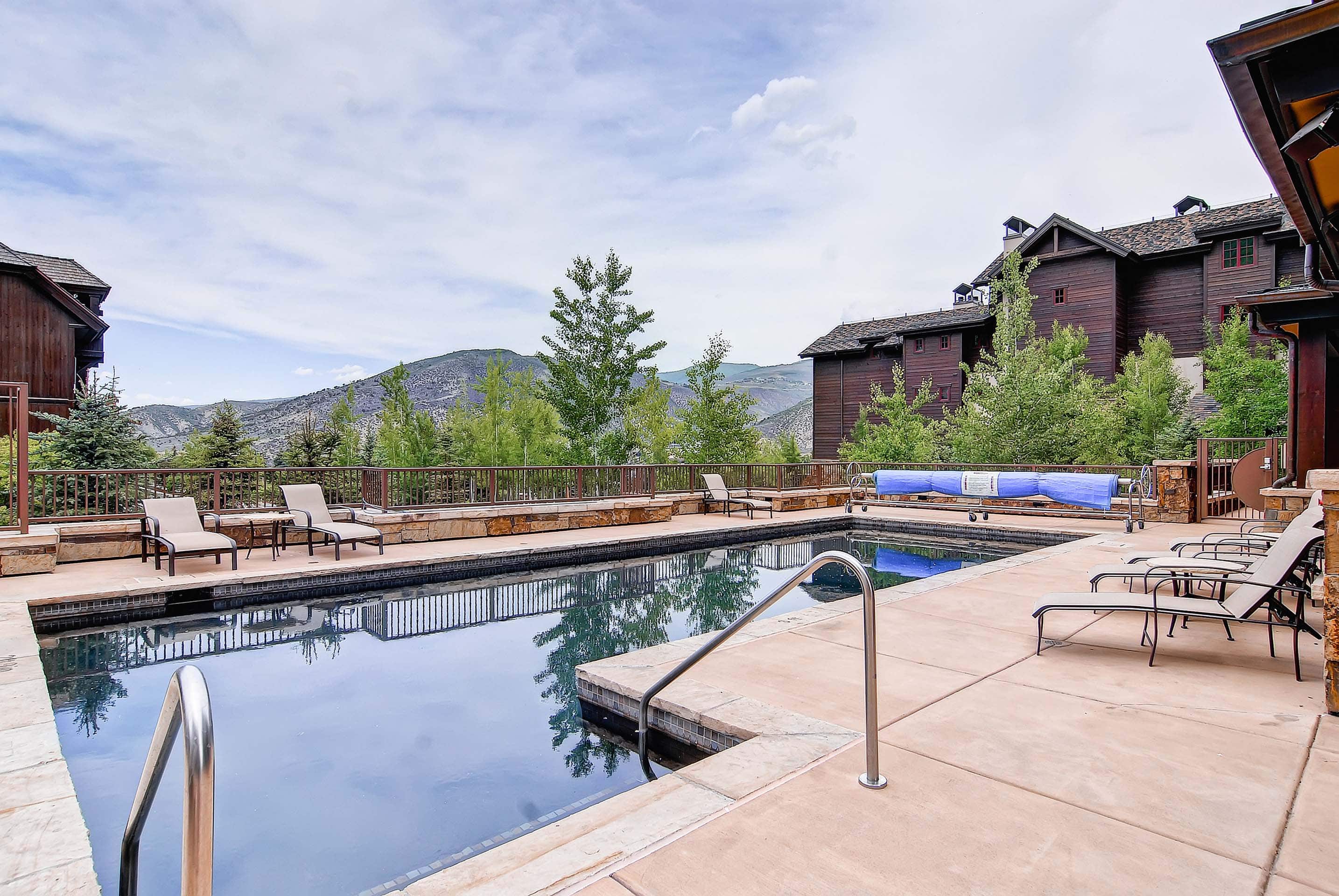 The image shows an inviting outdoor pool area with lounge chairs arranged on the surrounding deck. The pool's water reflects the sky and adjacent trees, creating a serene ambiance. A large, luxurious-looking building can be seen in the background, suggesting a resort or high-end residential complex.