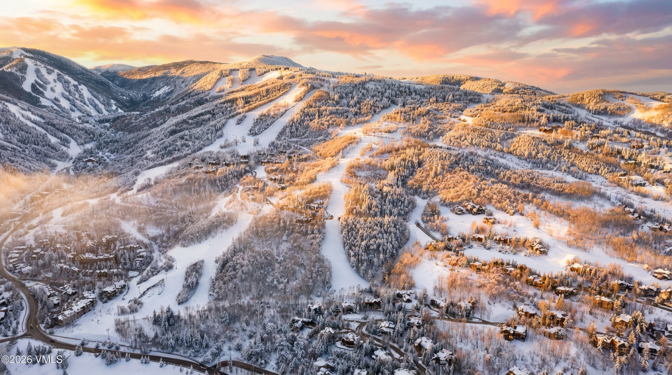 This is an aerial view showcasing the exterior of multiple mountain residences surrounded by snow-covered trees and ski slopes. The image highlights the properties idyllic location within the mountainous landscape, emphasizing access to skiing. The golden hour lighting adds warmth and enhances the appeal of the residences nestled among the trees.