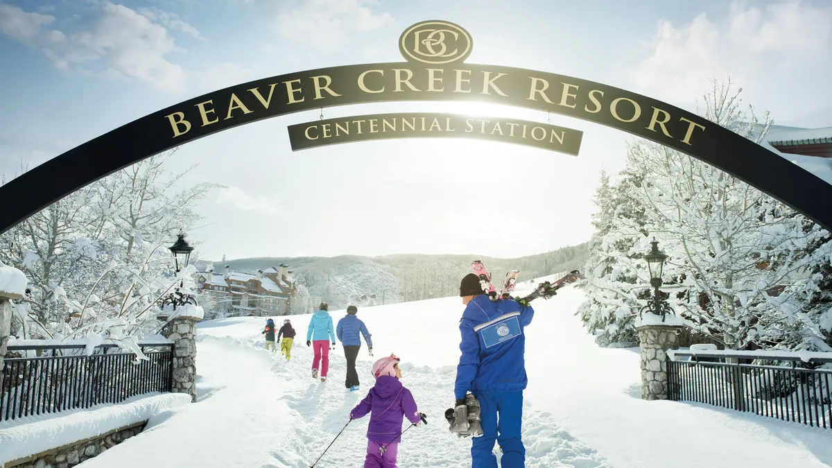 The image captures the entryway to Beaver Creek Resort in a snowy landscape. A group of people, including children, are walking toward the resort under the overhead sign. The scene evokes a sense of arrival and winter recreation, ideal for showcasing the location for potential residents or vacationers.