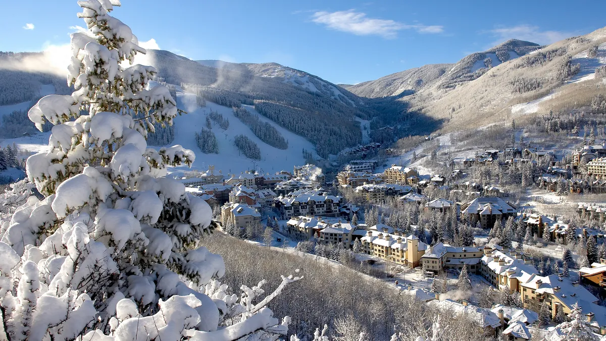 This aerial view showcases a picturesque mountain town blanketed in snow. The buildings are nestled among snow-covered trees, evoking a cozy and inviting atmosphere. Ski slopes can be seen on the mountains in the background, indicating a prime location for winter recreation.