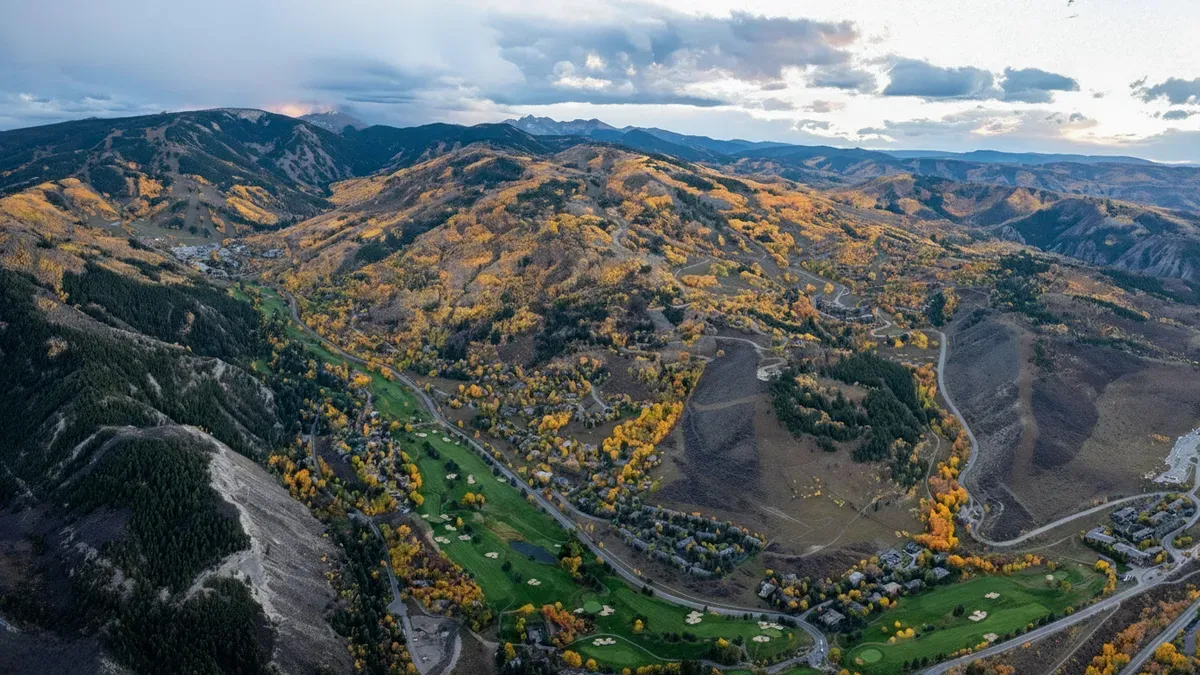 This aerial view captures a vast landscape encompassing mountains and a golf course community. The mountains are adorned with autumnal foliage, presenting a vibrant and picturesque scene. The golf course weaves through the residential area, providing a recreational amenity within a serene setting.