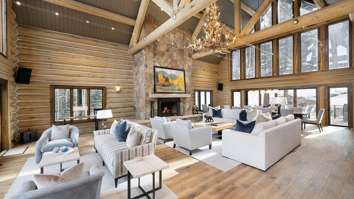 This is a spacious living room with a rustic yet elegant style. The room features log walls, a stone fireplace, and large windows that provide plenty of natural light. The antler chandelier adds a unique touch, complementing the overall lodge-like atmosphere.