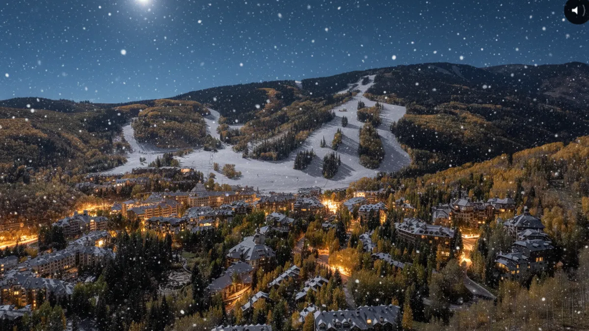 This aerial view showcases a luxurious mountain resort town covered in a fresh blanket of snow. The scene features numerous chalet-style buildings nestled among evergreen trees, with ski slopes visible on the mountainside in the background. The overall impression is one of a high-end winter destination and pristine natural beauty.