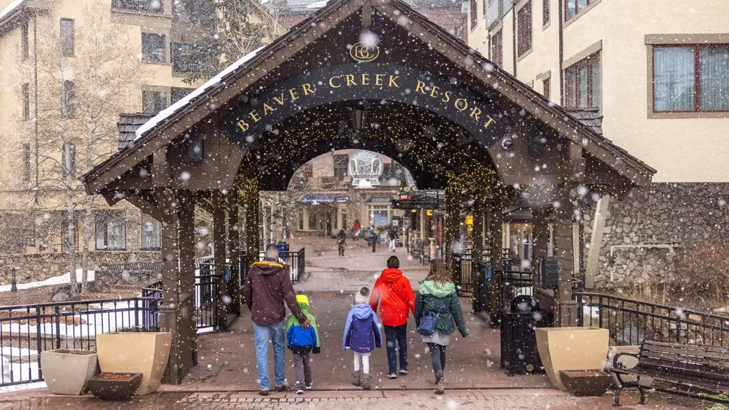 This picture shows a view of Beaver Creek Resort under a covered walkway. People are walking into the resort. There is snow falling, creating a winter wonderland atmosphere. 